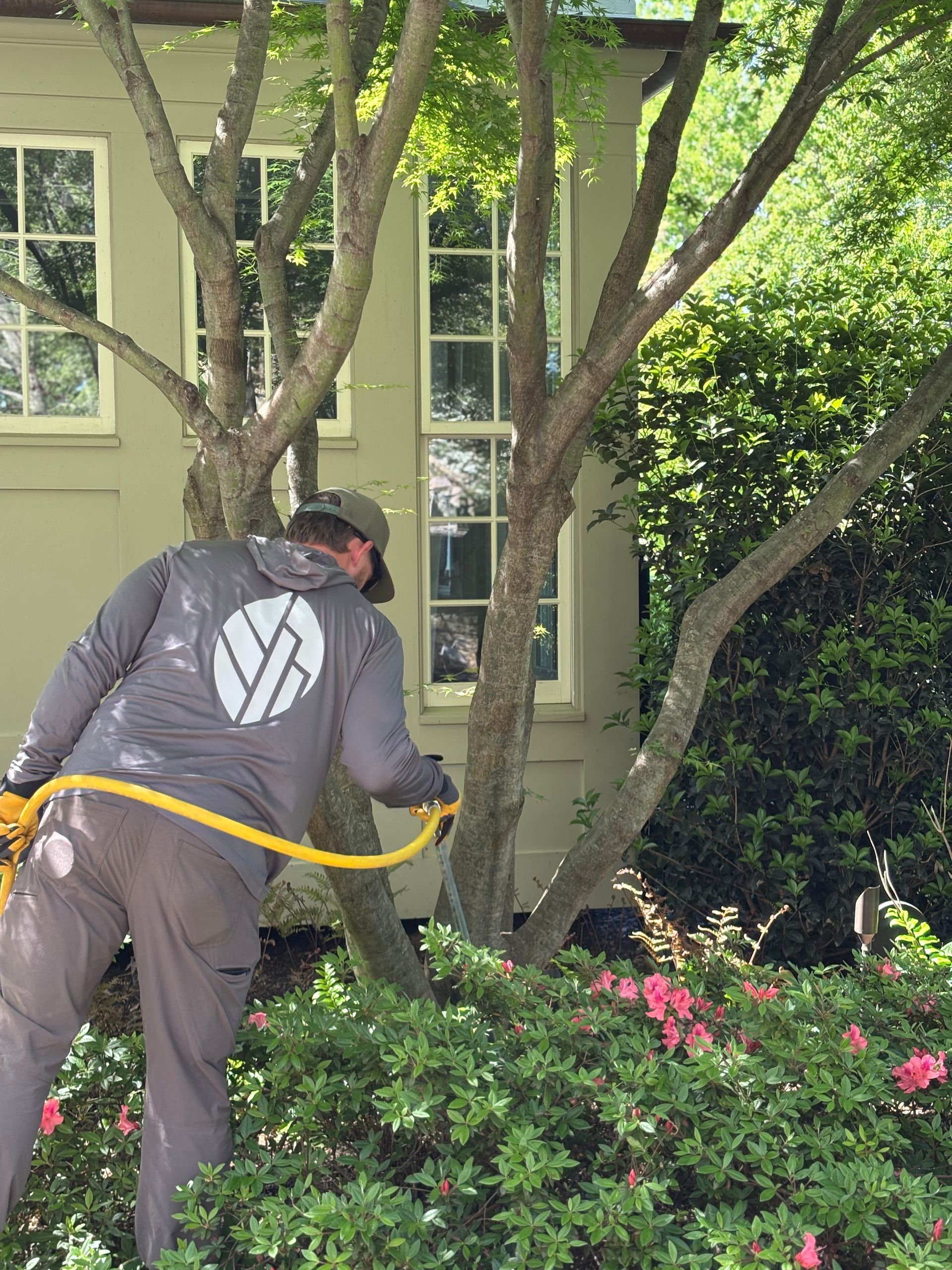 Worker trimming bushes beside a house, wearing a gray jacket and black hat, with a yellow hose in front.