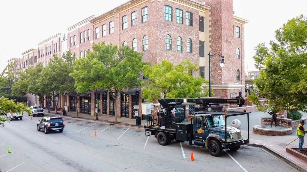 City street with brick buildings, parked utility truck, cones, trees, and a few pedestrians on the sidewalk