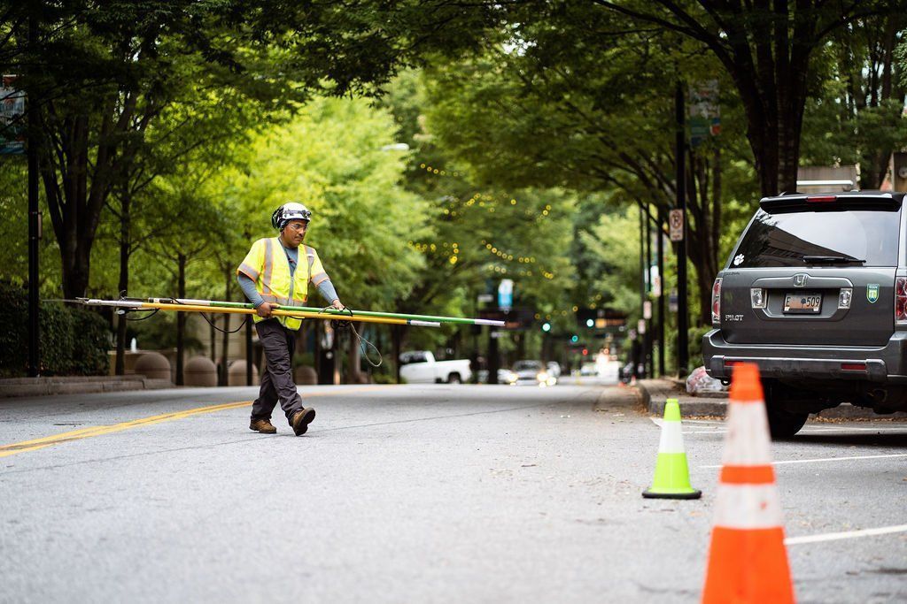 Traffic worker in yellow vest carrying a barrier on a tree-lined road beside parked cars and cones