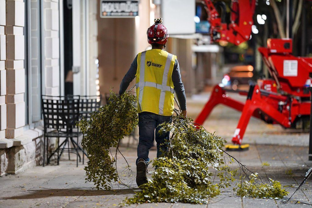 Worker in yellow vest clearing tree branches from a street near red construction equipment.