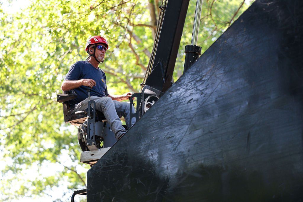 Worker in red helmet on a lift, painting a large black structure outdoors among trees.