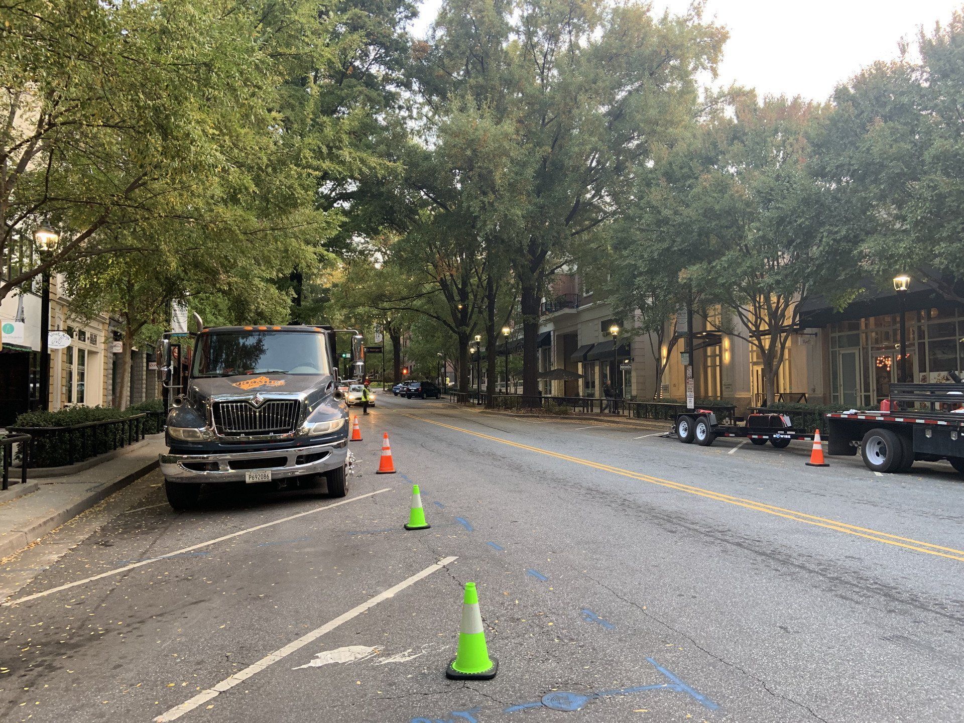 Tree-lined city street with a parked truck, orange cones, and green markers guiding traffic lane closure
