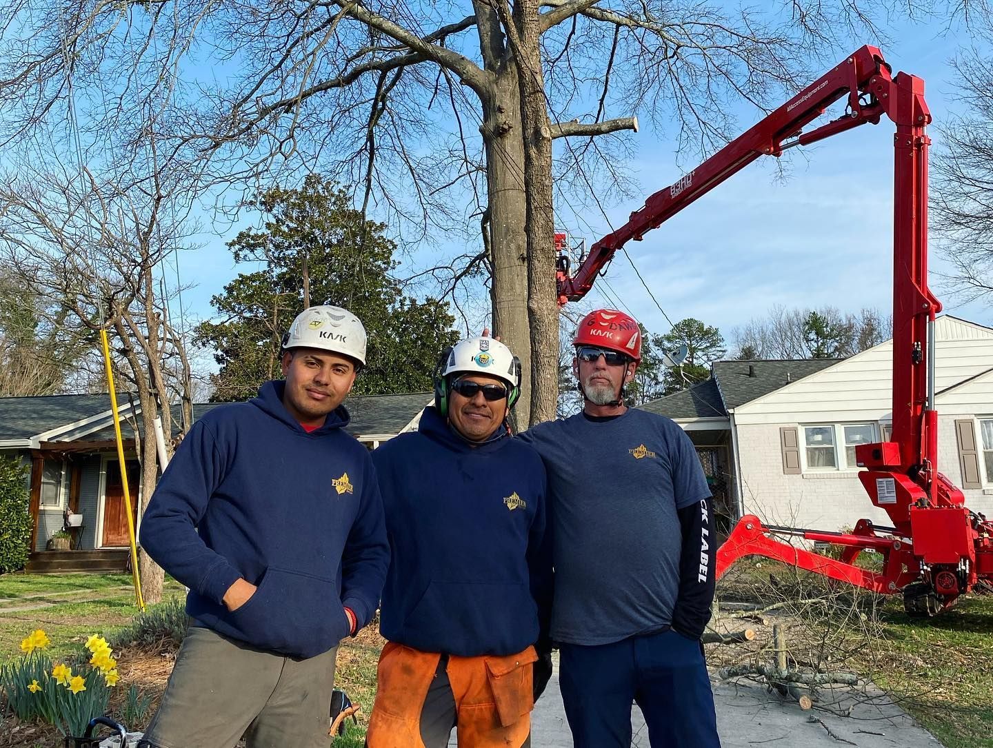 Three workers in hard hats pose outdoors beside a red boom lift and a large tree.