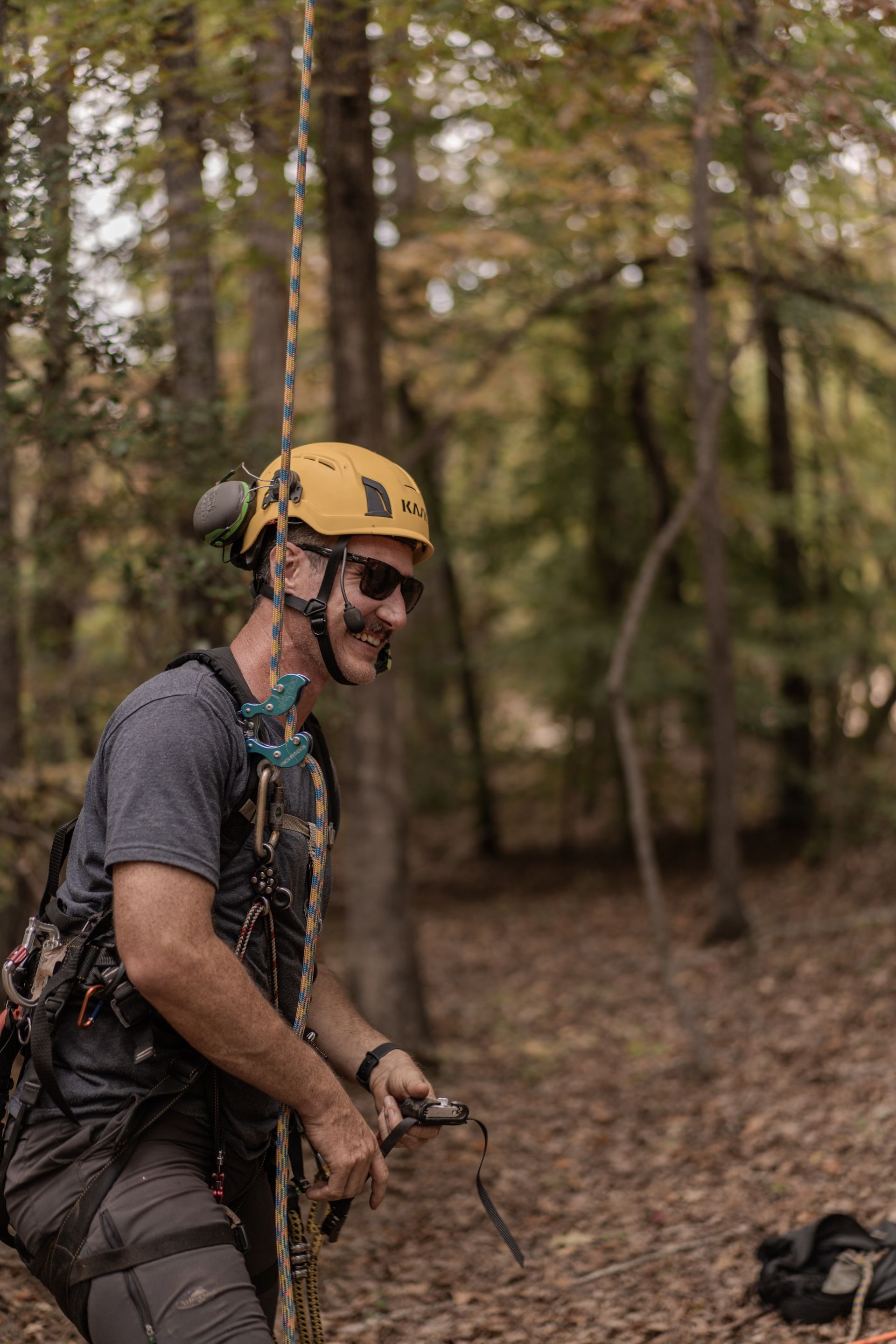 Climber in a yellow helmet and harness smiling in a wooded area, holding a rope.