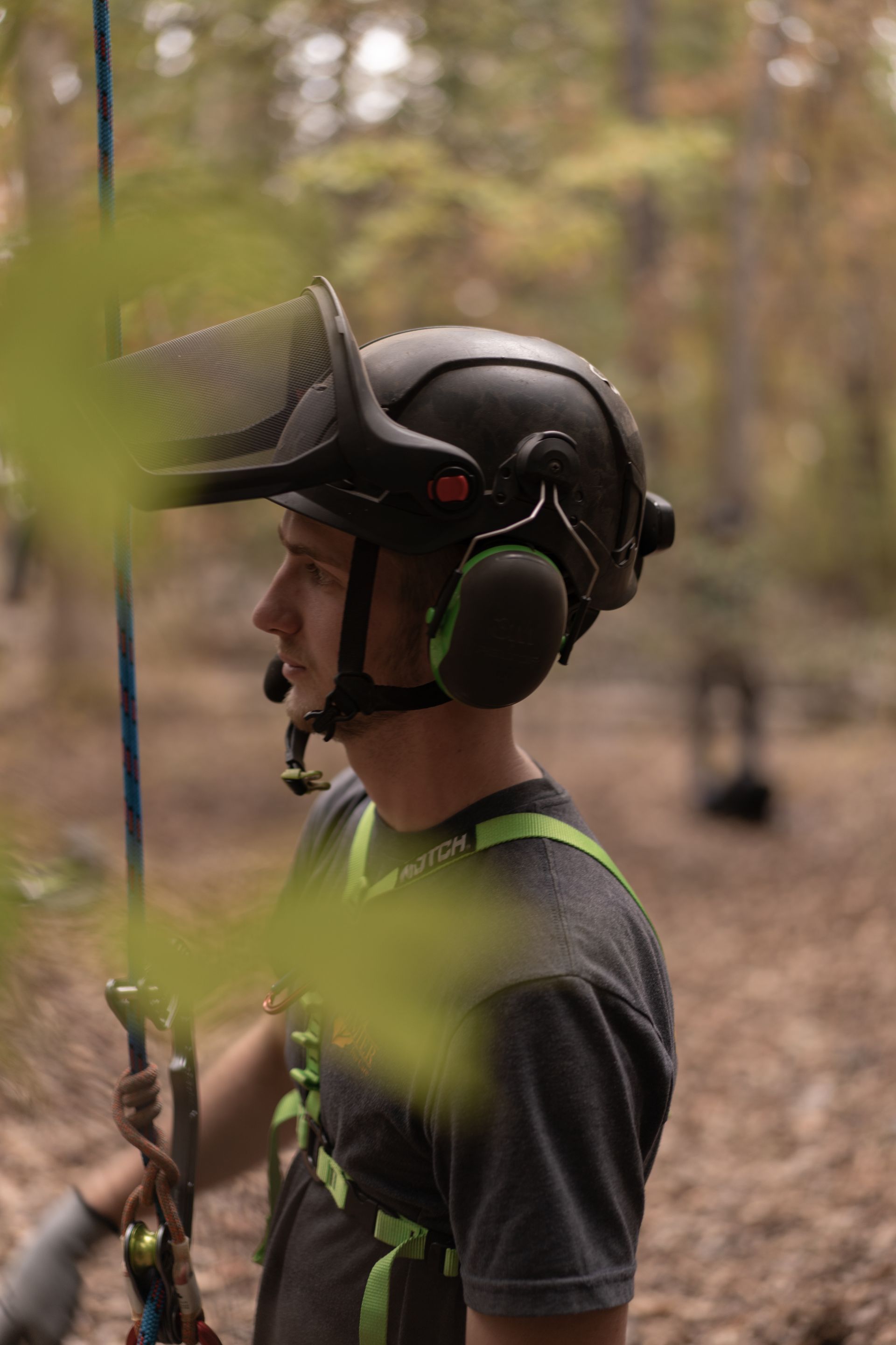 Person wearing a helmet and headset, standing in a wooded area with climbing gear.