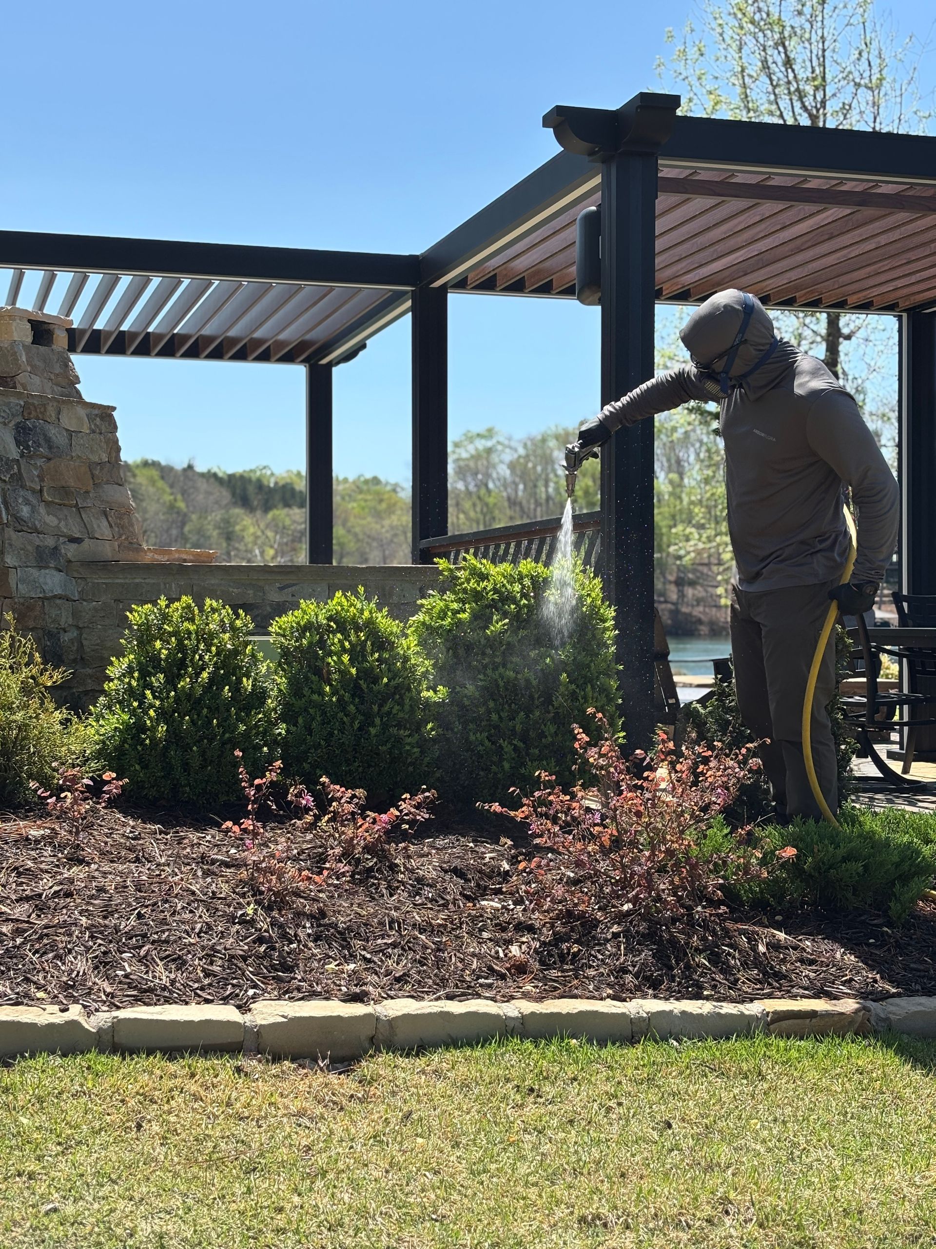 Person watering shrubs in a landscaped garden beside a pergola on a sunny day