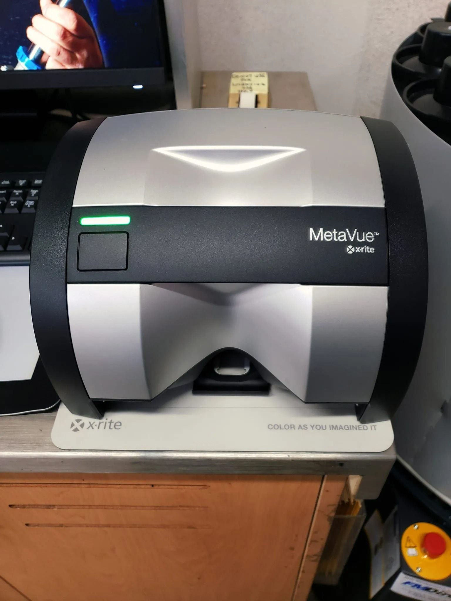 Silver and black MetaVue VS320 color measurement instrument on a table, with a green light on.
