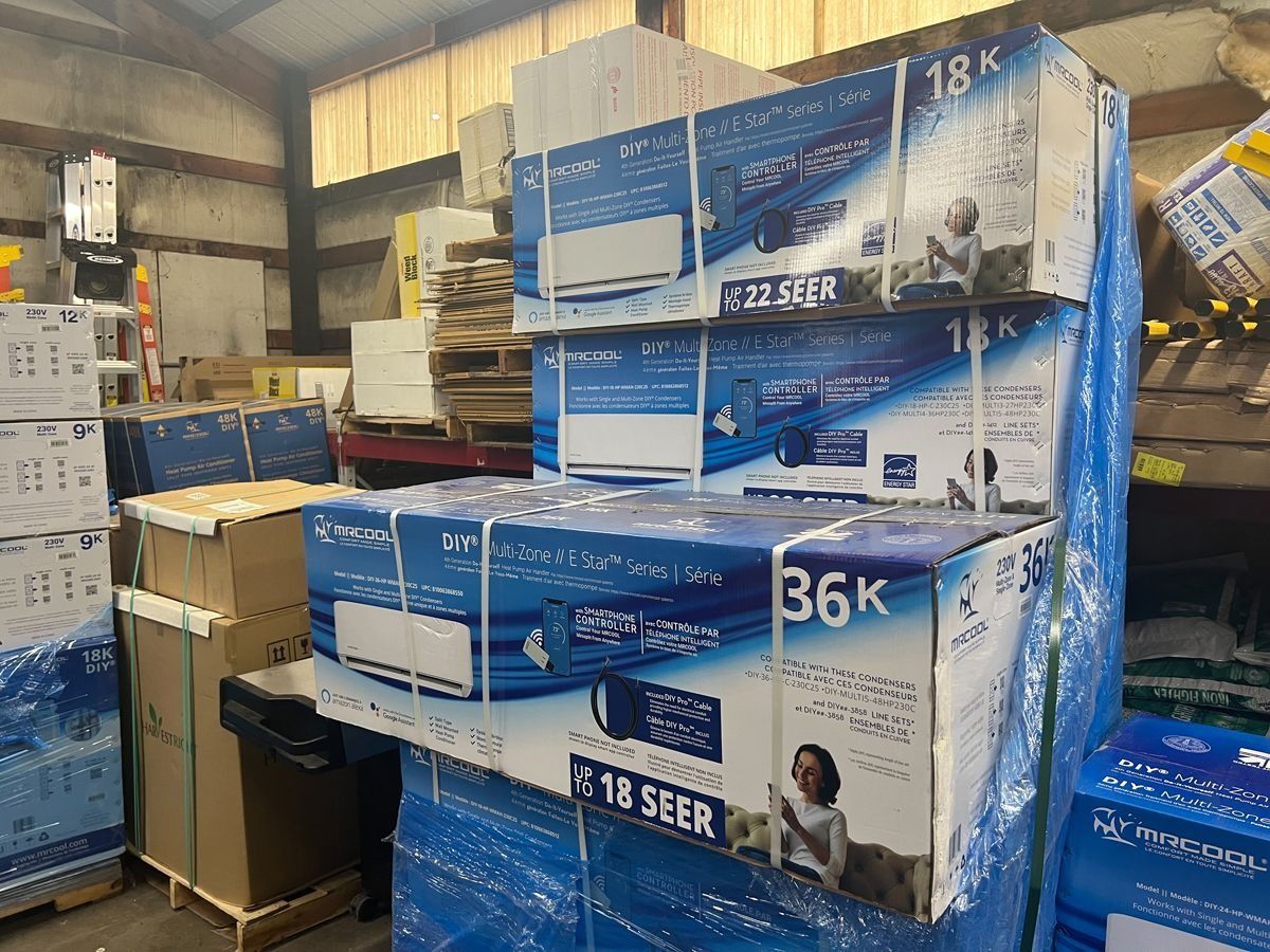 Pallet of stacked air conditioner boxes in a warehouse. Blue and white boxes, plastic wrap, and cardboard visible.
