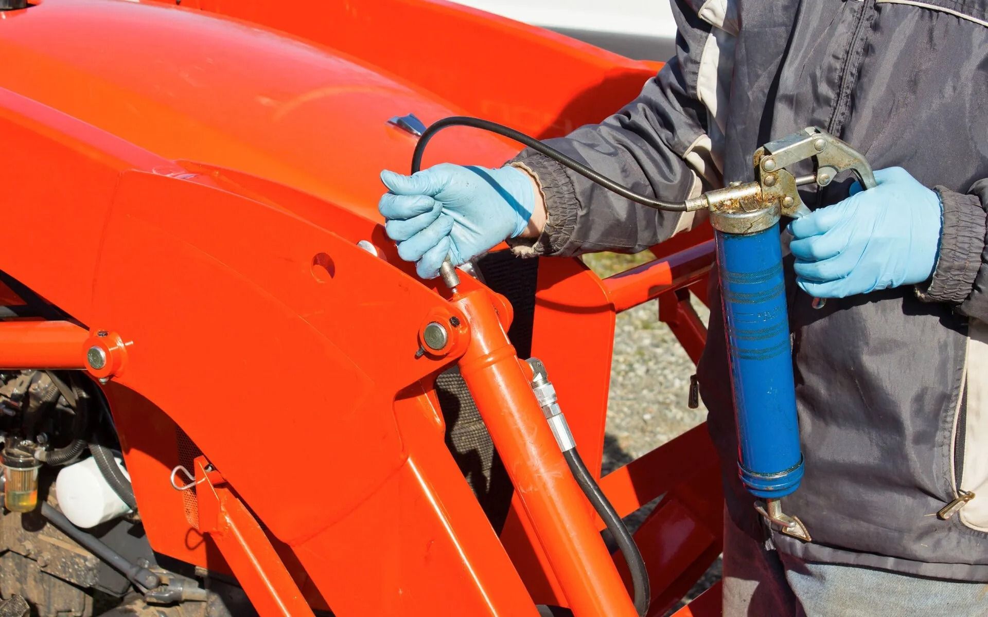 Person greasing a tractor with a blue grease gun on an orange tractor.