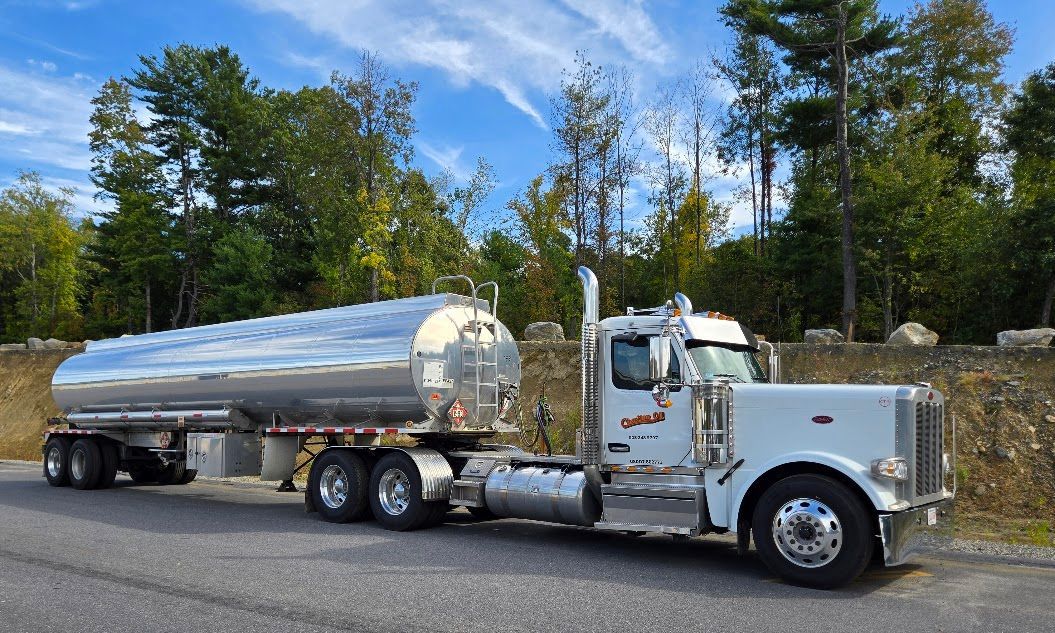 White tanker truck on a road with a stainless steel tank, trees and a blue sky in the background.