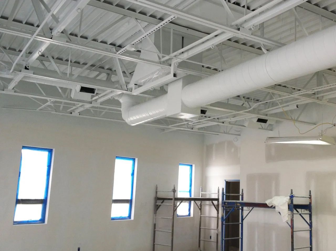 Interior of a room under construction; white painted ceiling, ductwork, and partially finished walls; windows with blue taped edges.
