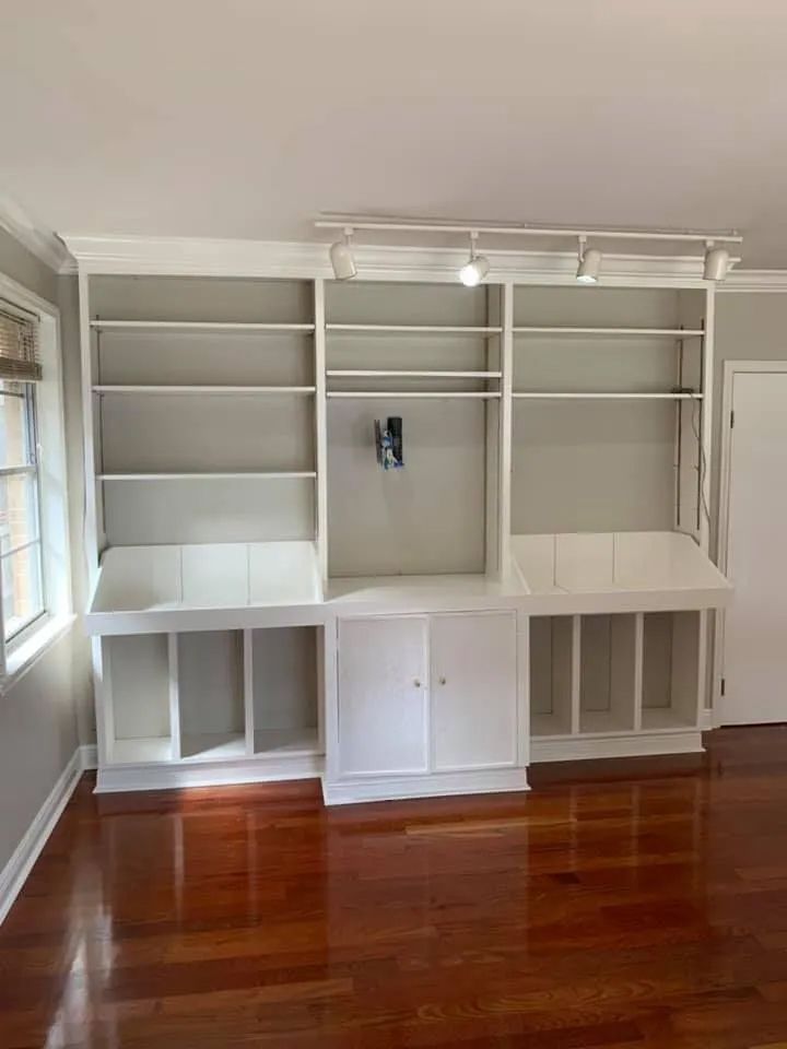 Built-in white shelving unit on a hardwood floor; includes upper shelves, a middle space for a TV, and lower cabinets.