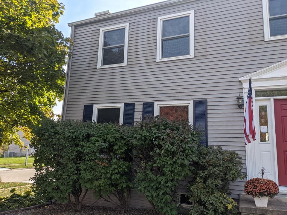 Two-story gray house with blue shutters, windows, and a red door. An American flag hangs to the side, with green bushes in front.
