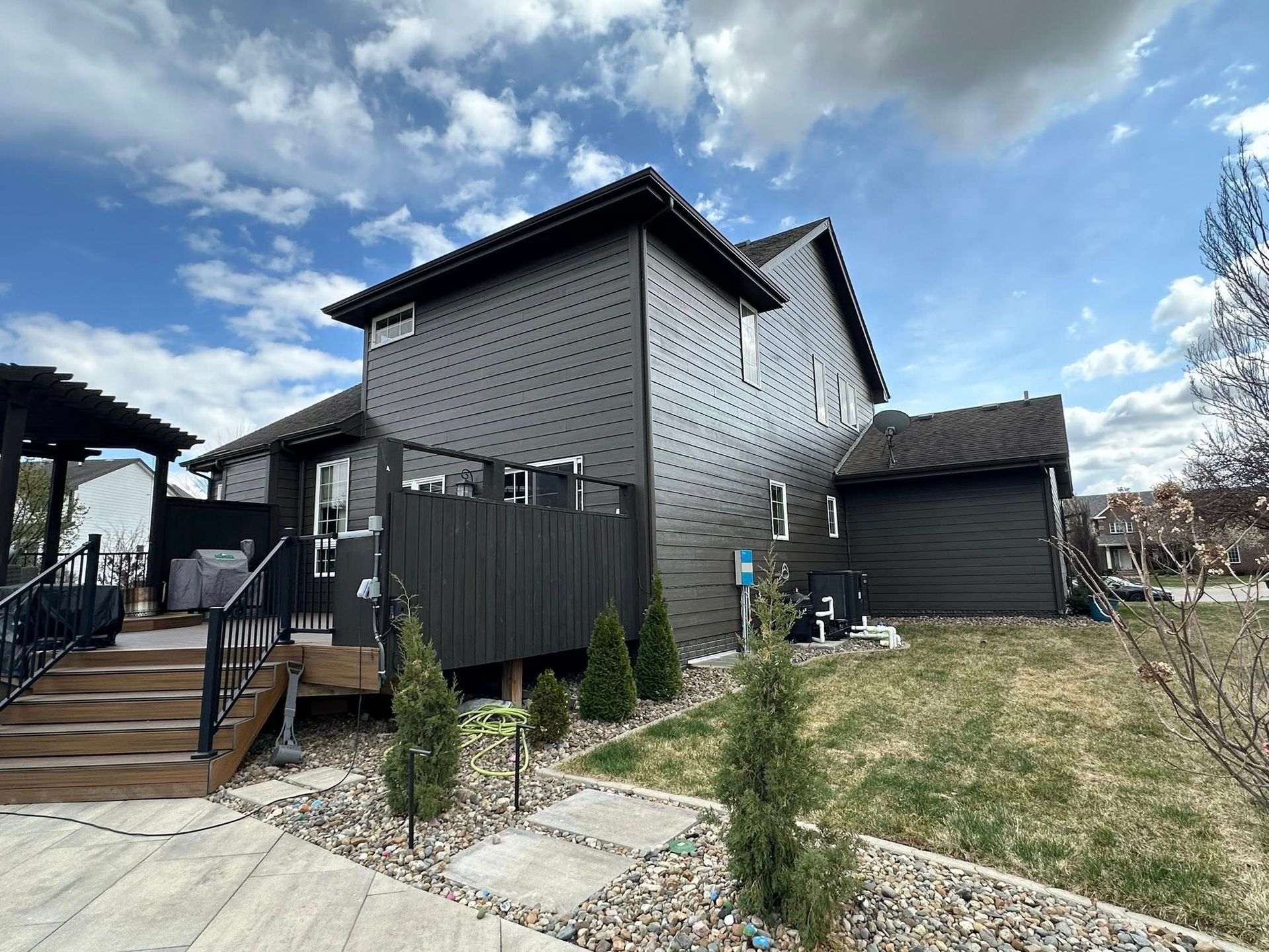 Dark gray house with black siding, deck, and landscaping under a cloudy blue sky.