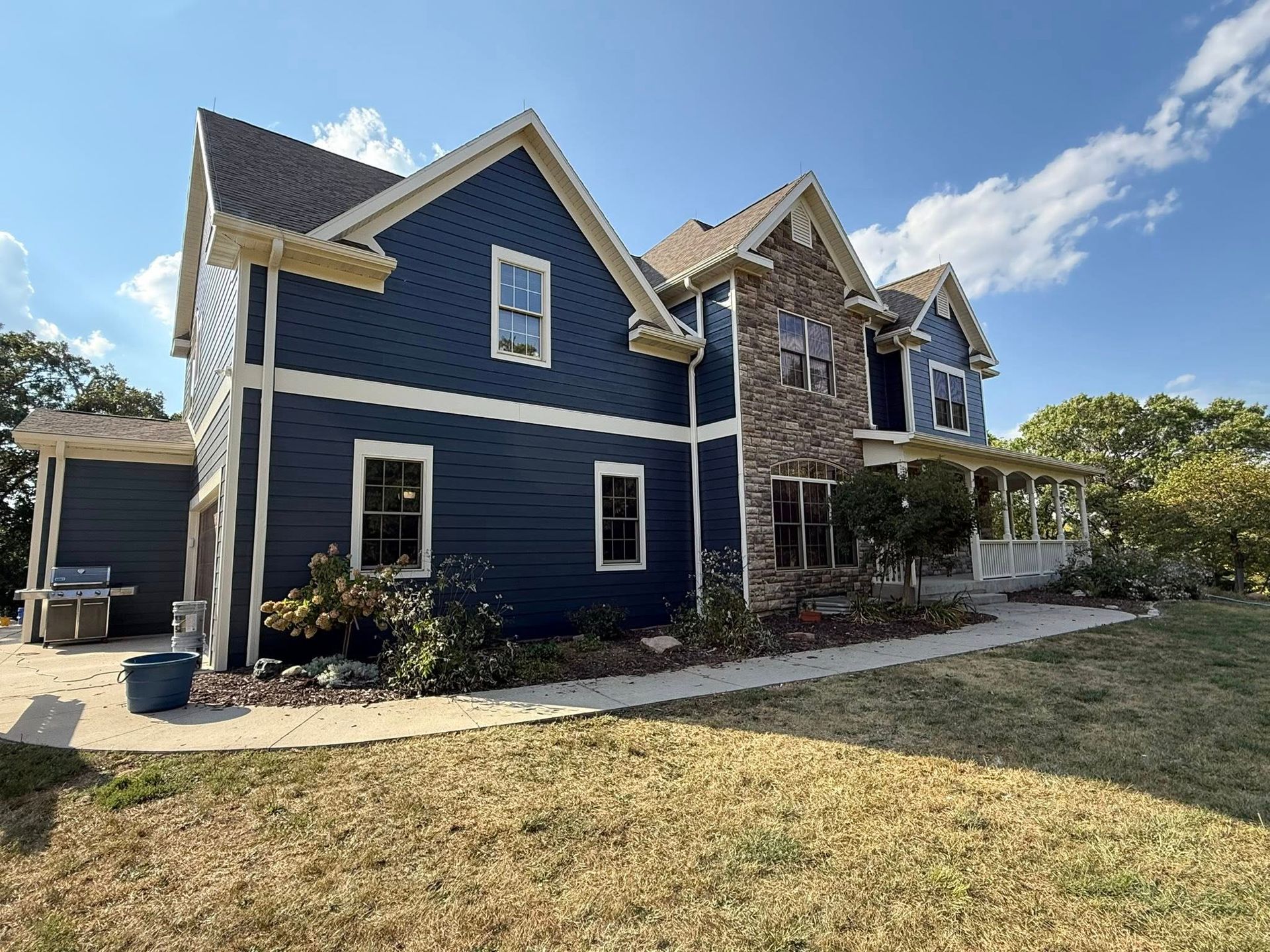 Two-story blue house with stone facade, white trim, and a wraparound porch on a sunny day.