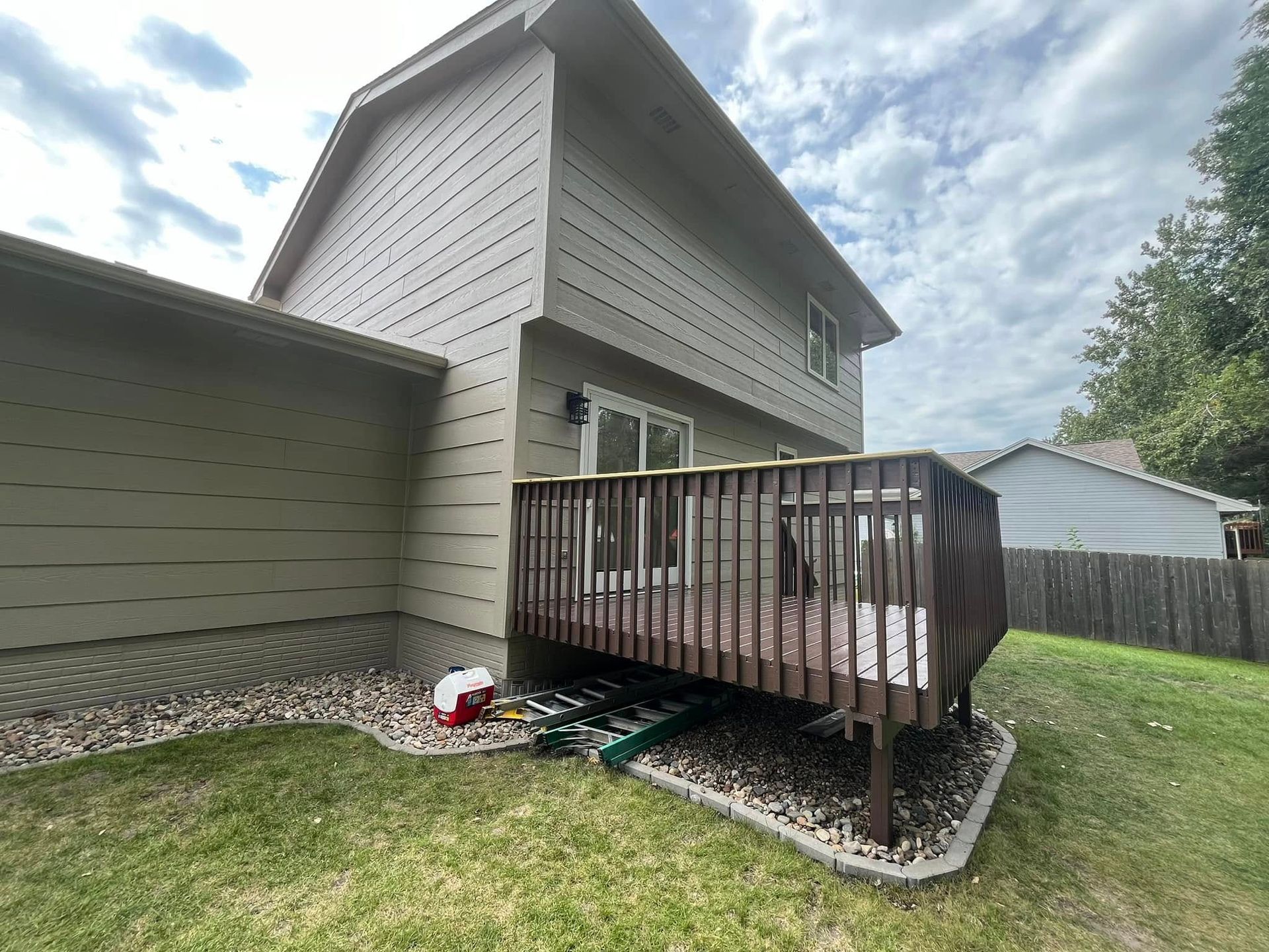 Two-story house with a wooden deck. Tan siding, brown deck, green grass, and cloudy sky.