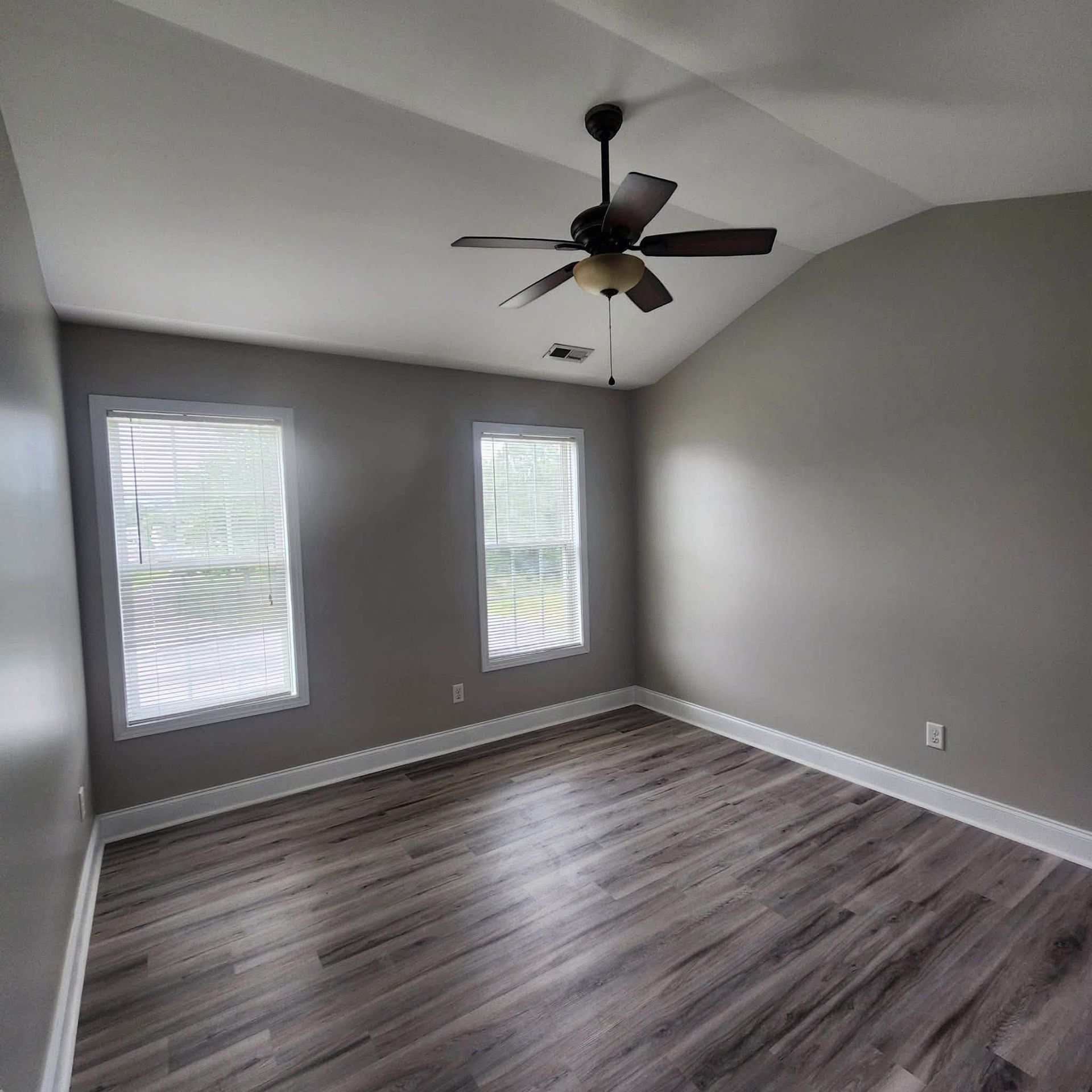 Empty gray bedroom with wood-look floor, three windows, and a ceiling fan.
