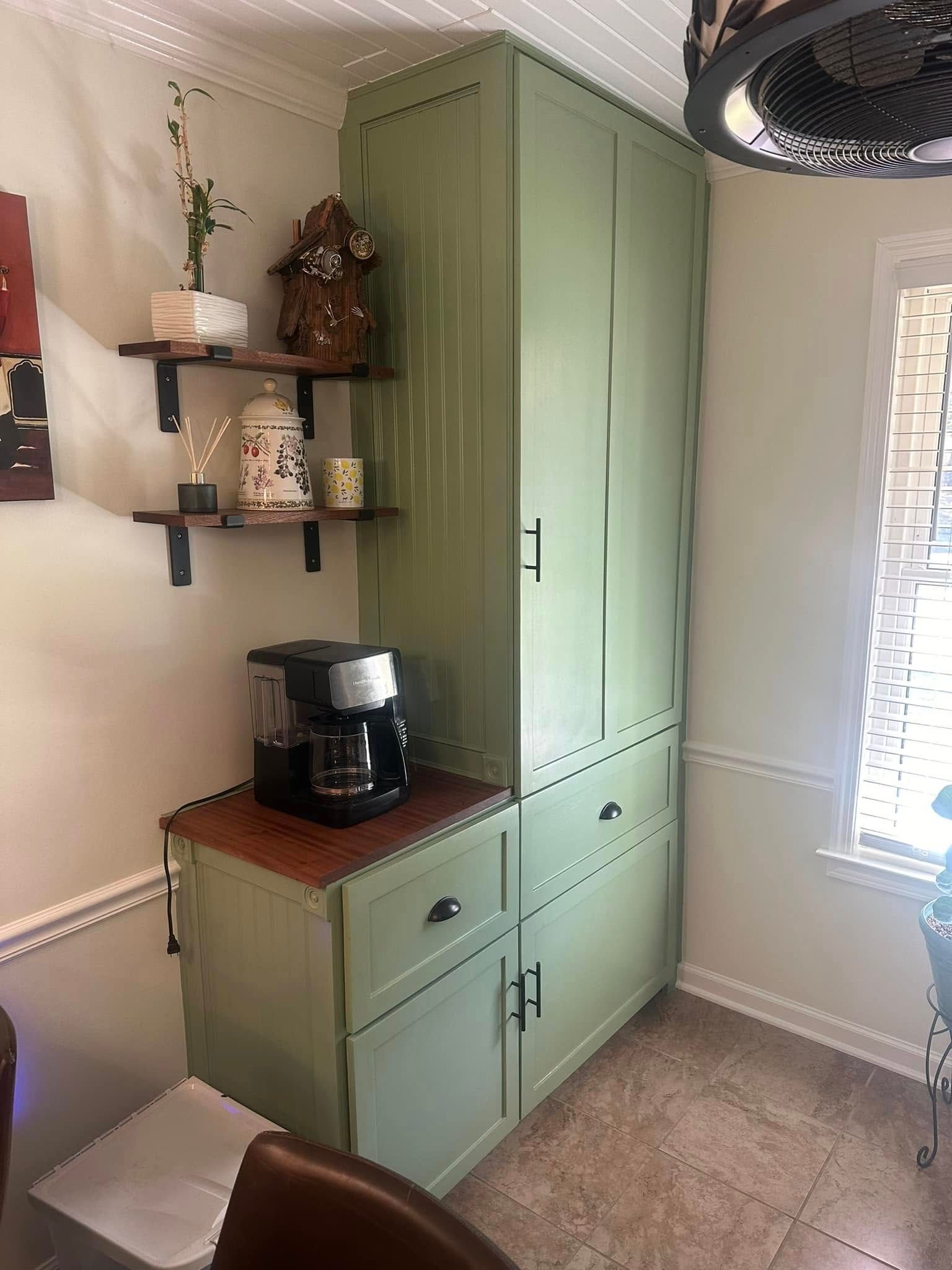 Green kitchen cabinet with a coffee maker on a wooden counter and wall shelves beside a window