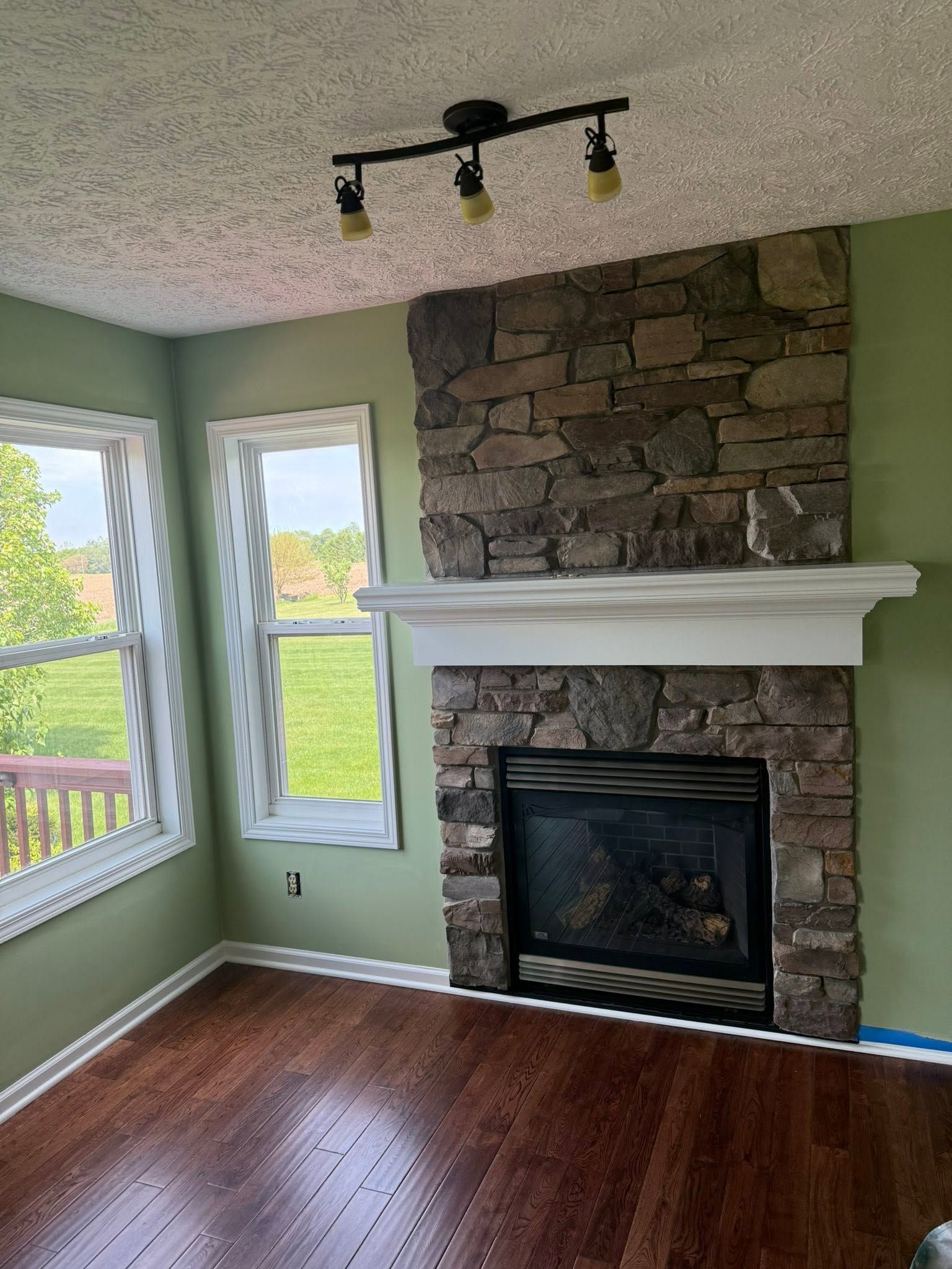 Empty room with hardwood floors, green walls, windows, and a stone fireplace with white mantel.