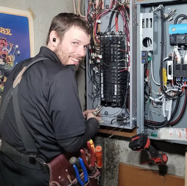 Electrician working on electrical panel, smiling. Tools and wires visible. Indoors, concrete wall.