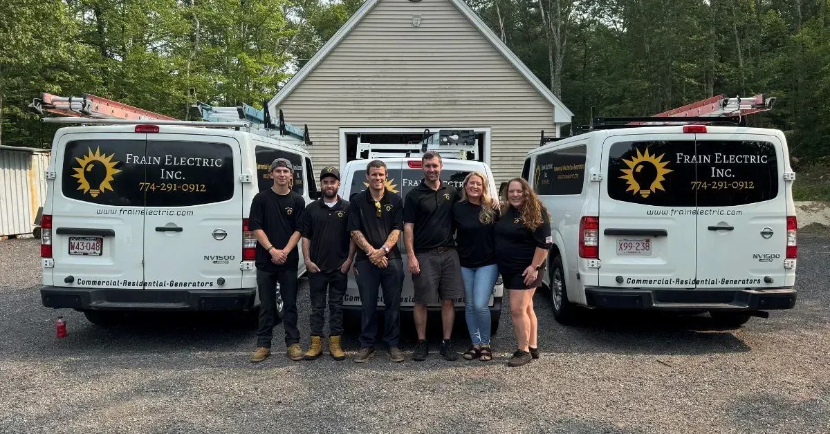 A group of people stand in front of two white vans with logos. They are in a gravel lot with a light-colored building.