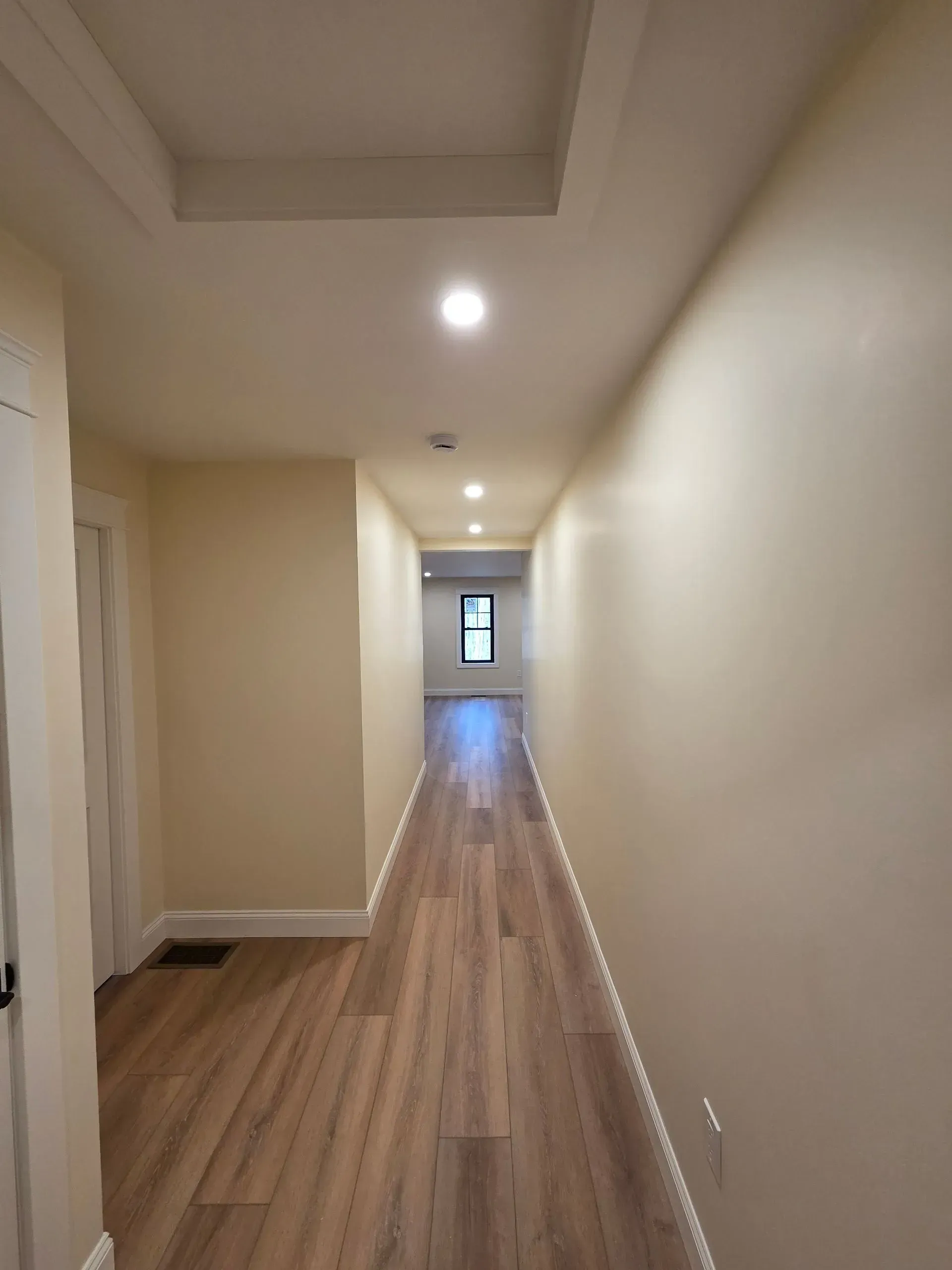 Long hallway with wooden floors, cream walls, and recessed ceiling lights. A door is on the left.