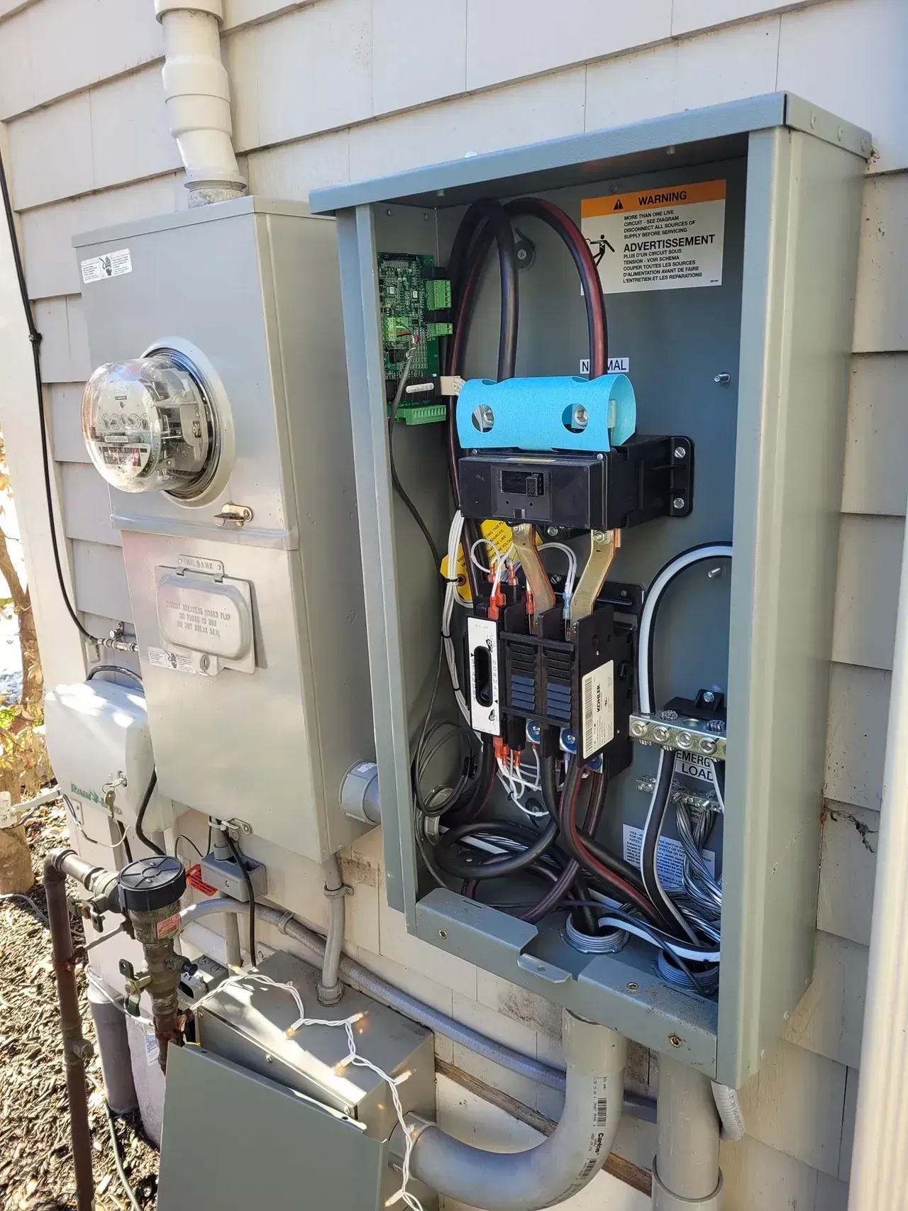 Outdoor electrical box with meter and exposed components on a house exterior. Wires, circuits, and a control unit visible.