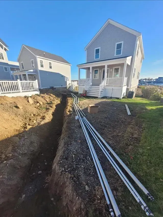 Narrow trench dug near houses, with white conduit pipes laid inside it.