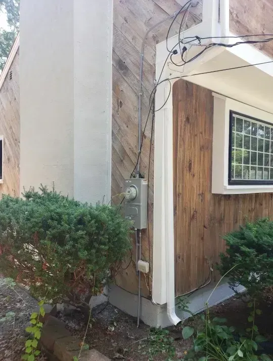 Exterior view of a house with wooden siding, a window, and electrical components mounted on the wall.