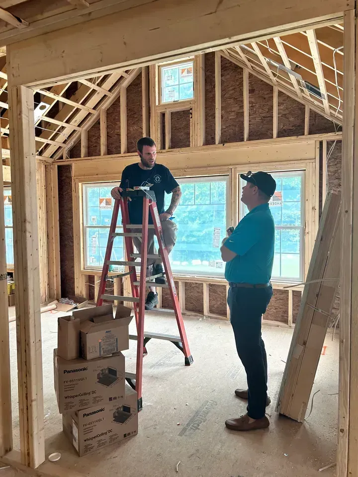 Two men inside a building under construction. One on a ladder, the other standing, observing. Boxes and lumber on the floor.
