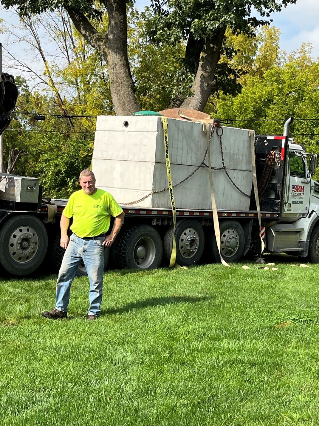 Truck hauling a large concrete block on grass beside a man in a neon shirt and trees