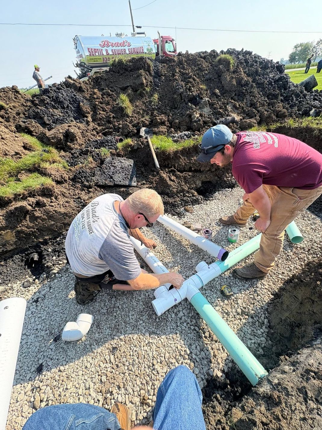 Two workers shoveling gravel into a trench beside a large dirt mound outdoors