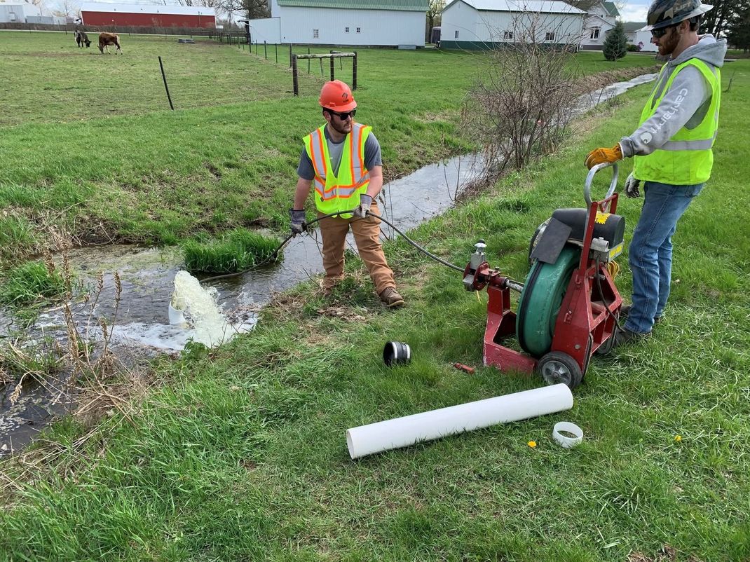 Workers repairing a grassy drainage ditch with a pipe and red pump machine