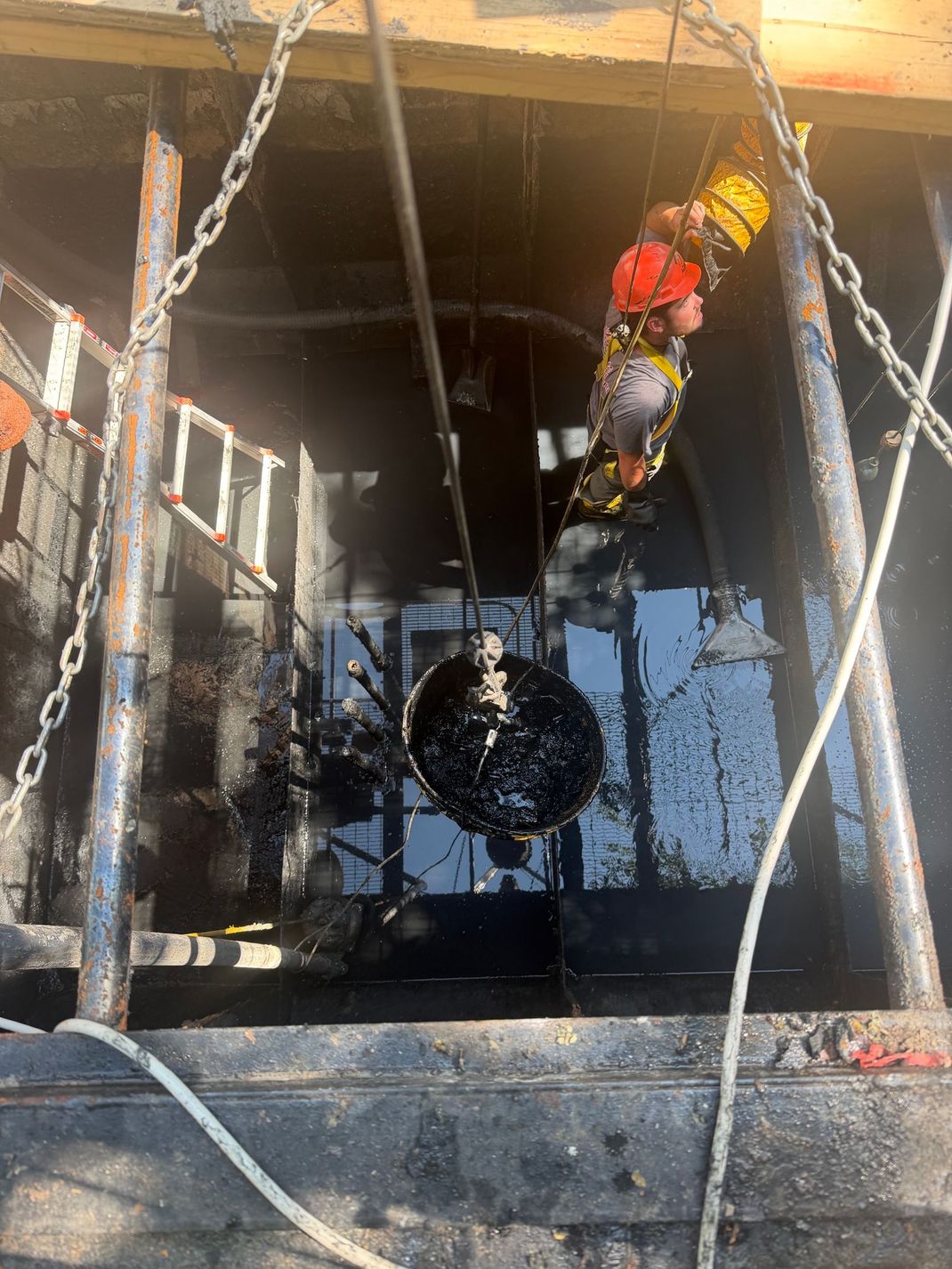 Worker in a hard hat descending into a dark manhole with ropes and ladder rails, wet interior visible