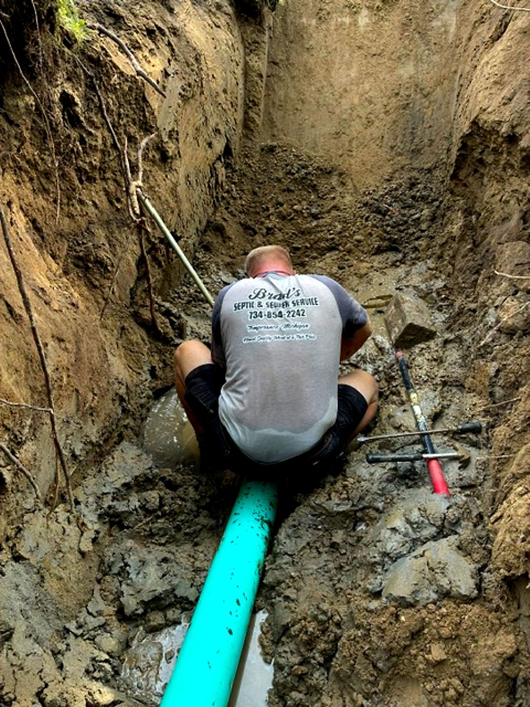 A worker crouches in a trench over a bright green pipe, using tools beside muddy earth walls.