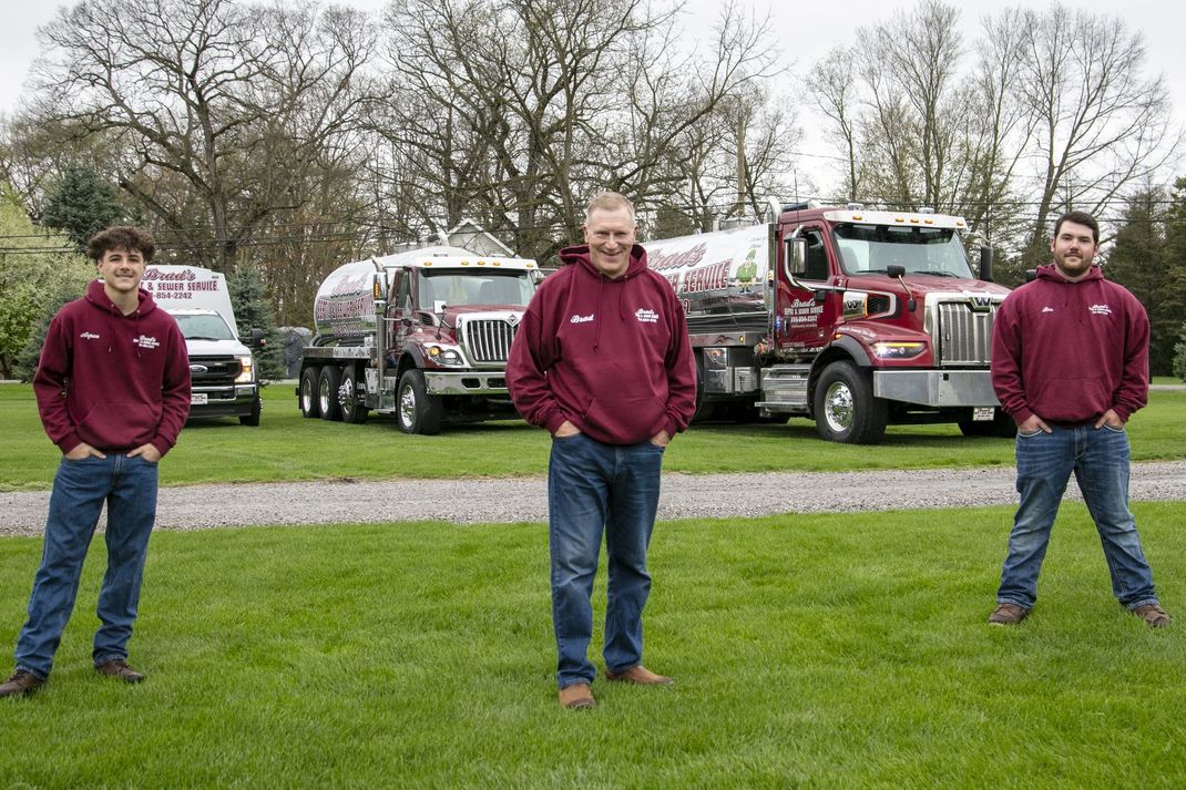 Three people in maroon hoodies stand on grass in front of trucks under cloudy trees.
