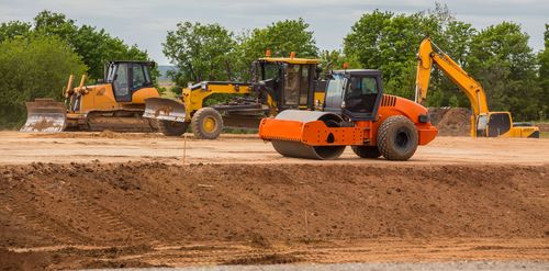 Orange construction vehicles grading a dirt site under cloudy skies