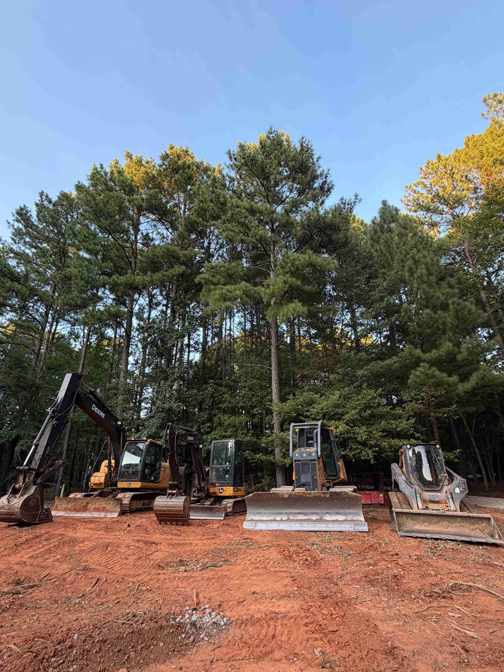 Excavators lined up on a dirt lot in front of tall trees under a clear blue sky