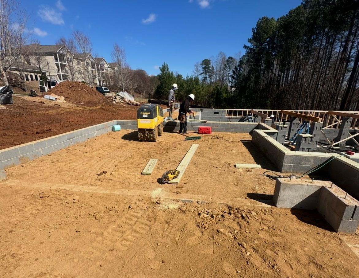 Construction site with yellow excavator grading sandy ground beside stone foundation walls under a clear blue sky