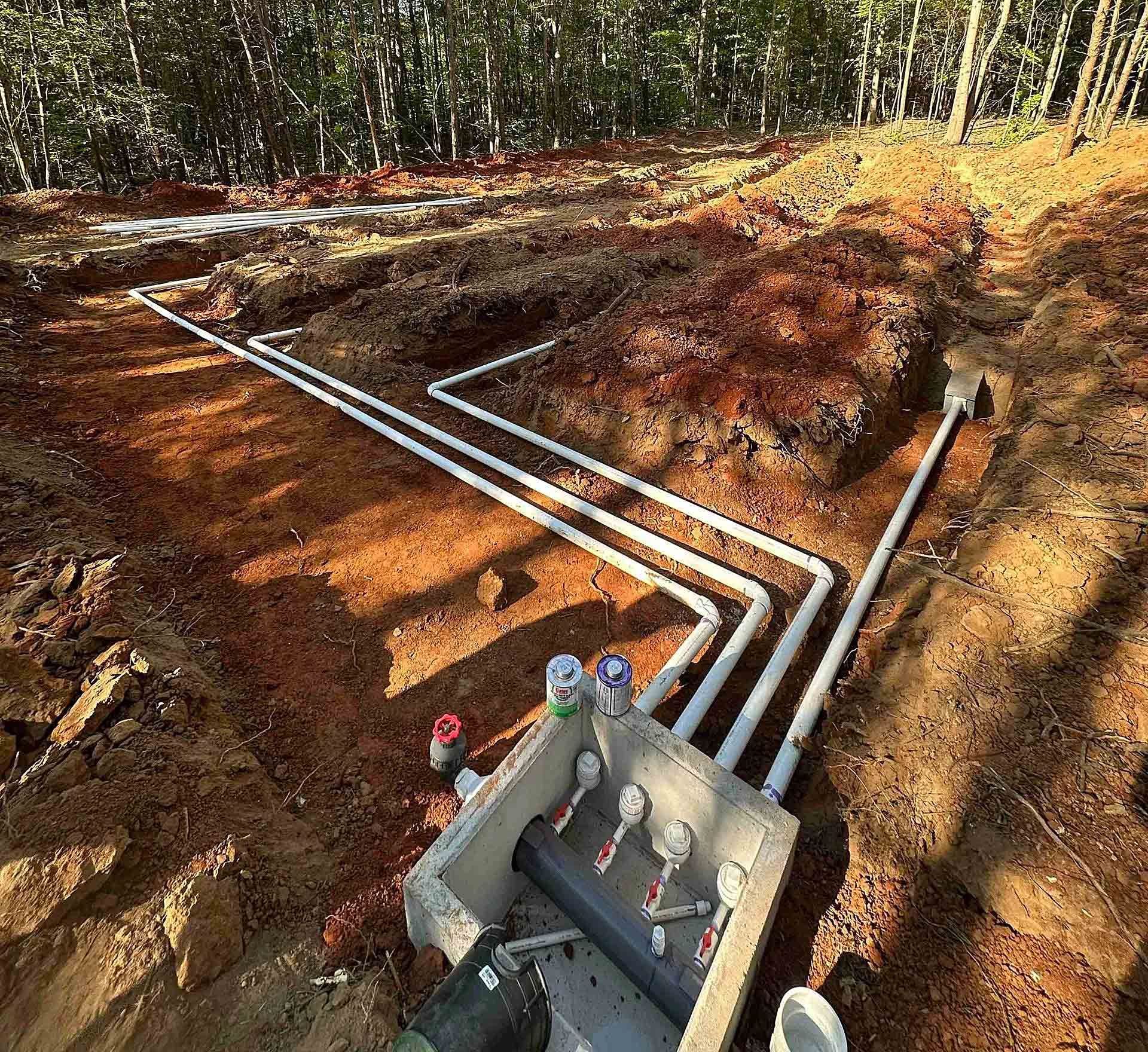 Underground utility trench with white pipes and a junction box on a wooded construction site.