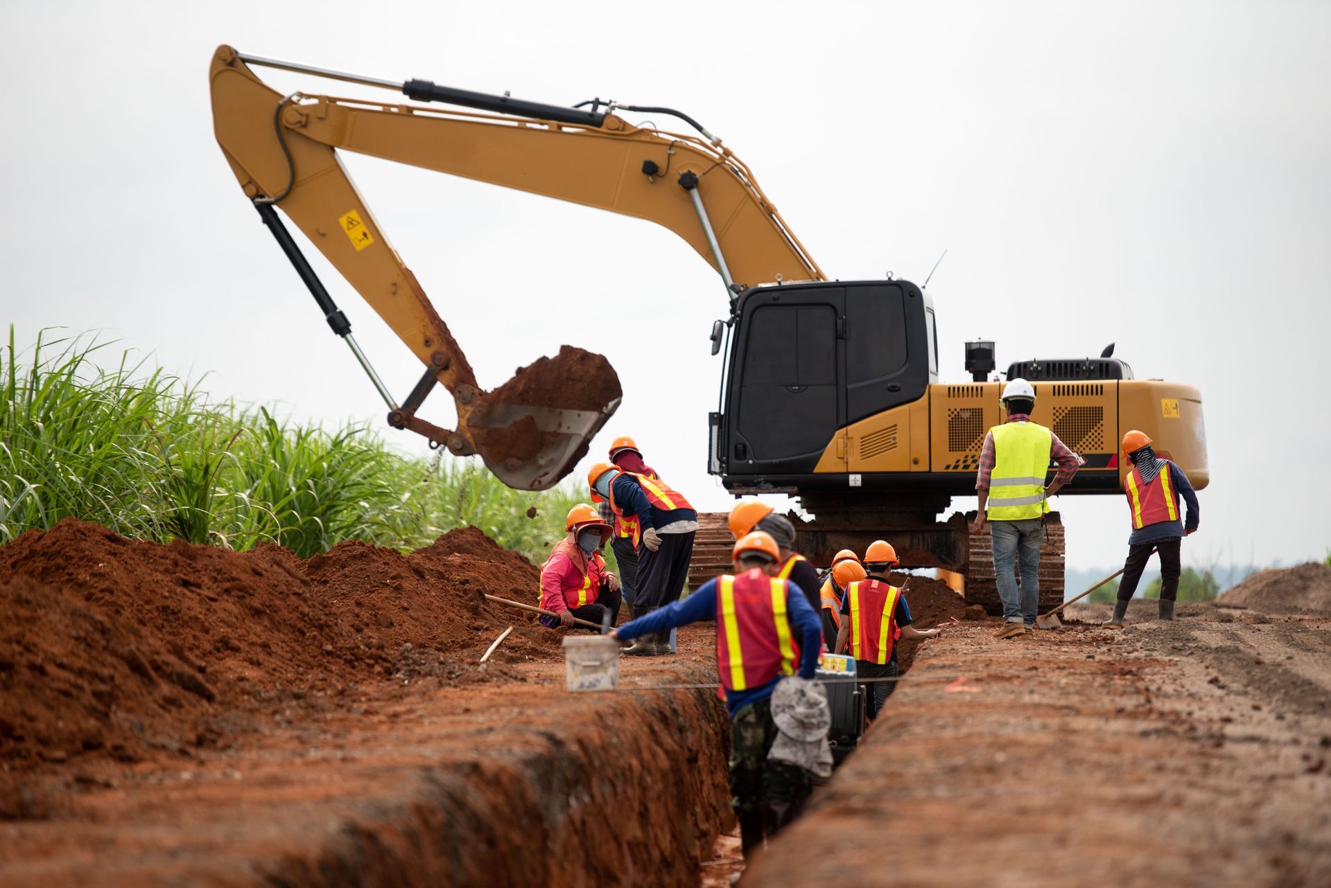 Construction crew works beside an excavator digging a trench on a dirt road.