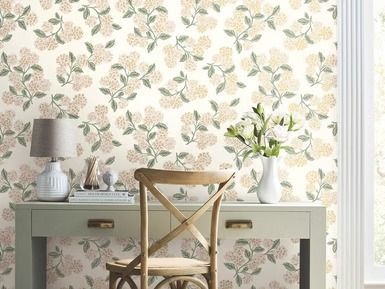 Desk and chair in front of a wall with floral wallpaper. A lamp, books, and vase with flowers sit on the desk.