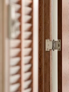 Close-up of a silver hinge attached to a brown wooden shutter with angled slats.