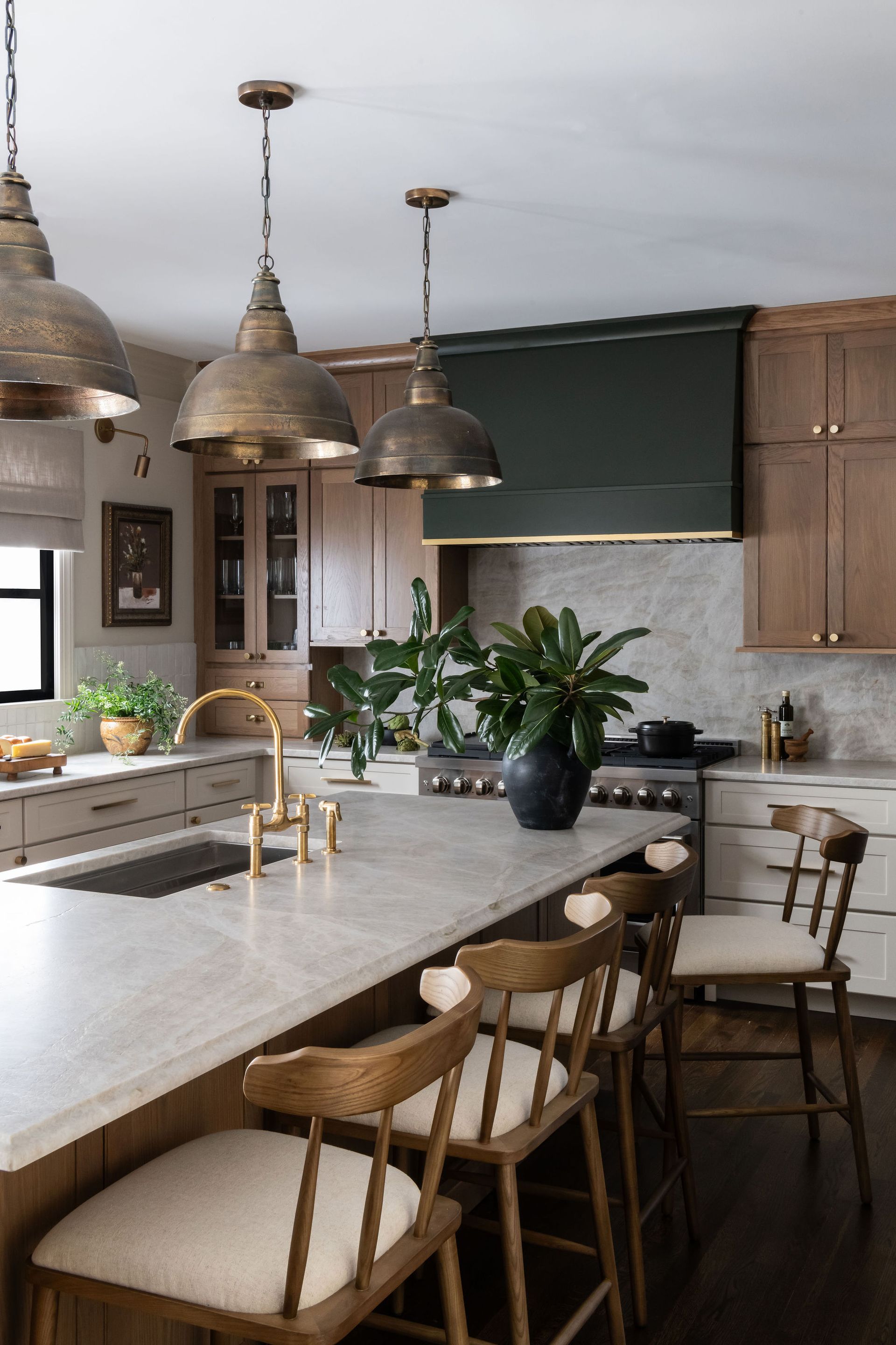 Kitchen with brass pendant lights, wooden stools, dark green range hood, and light countertops.