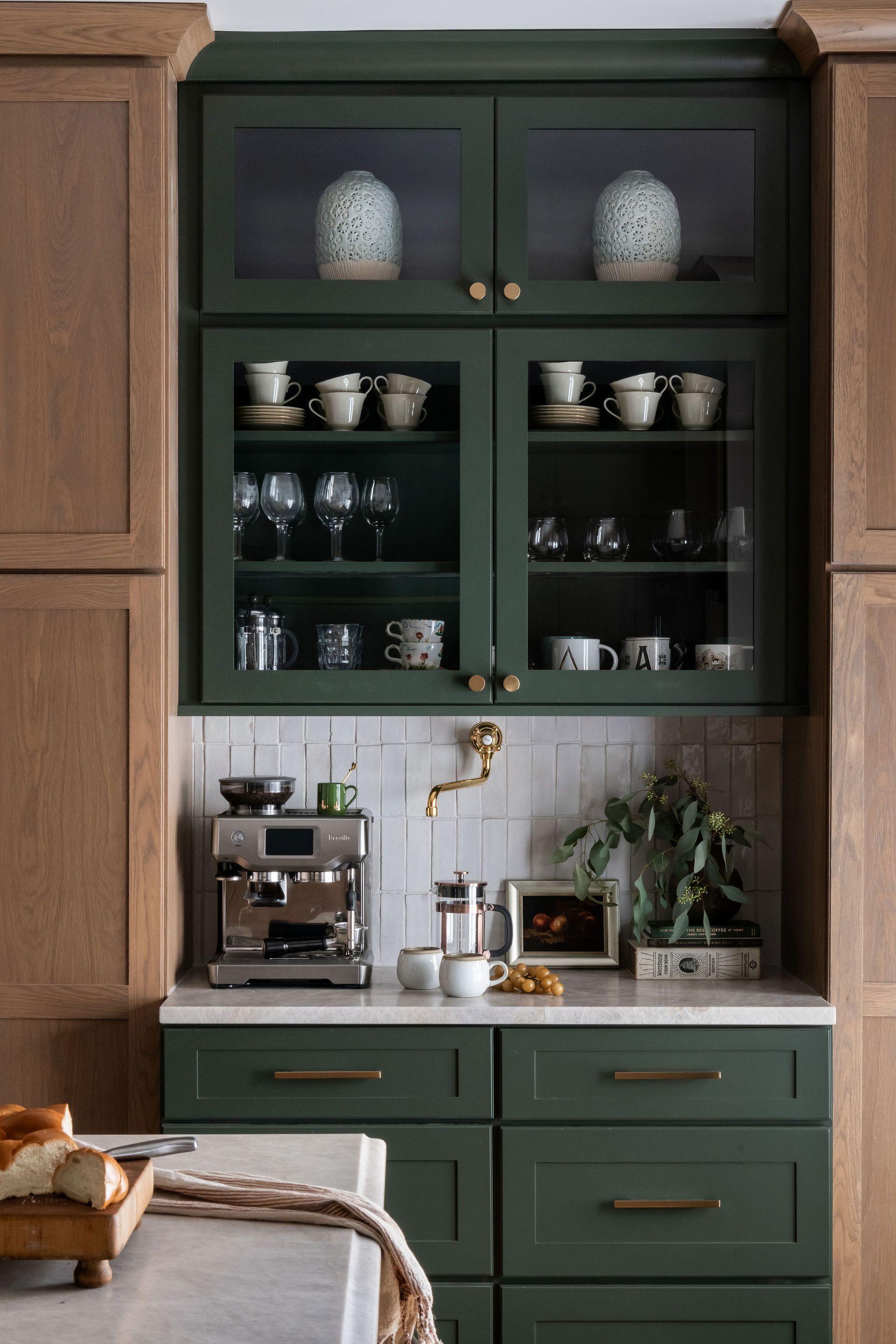 Green kitchen cabinets with glass doors displaying dishware. Espresso machine and sink on countertop.