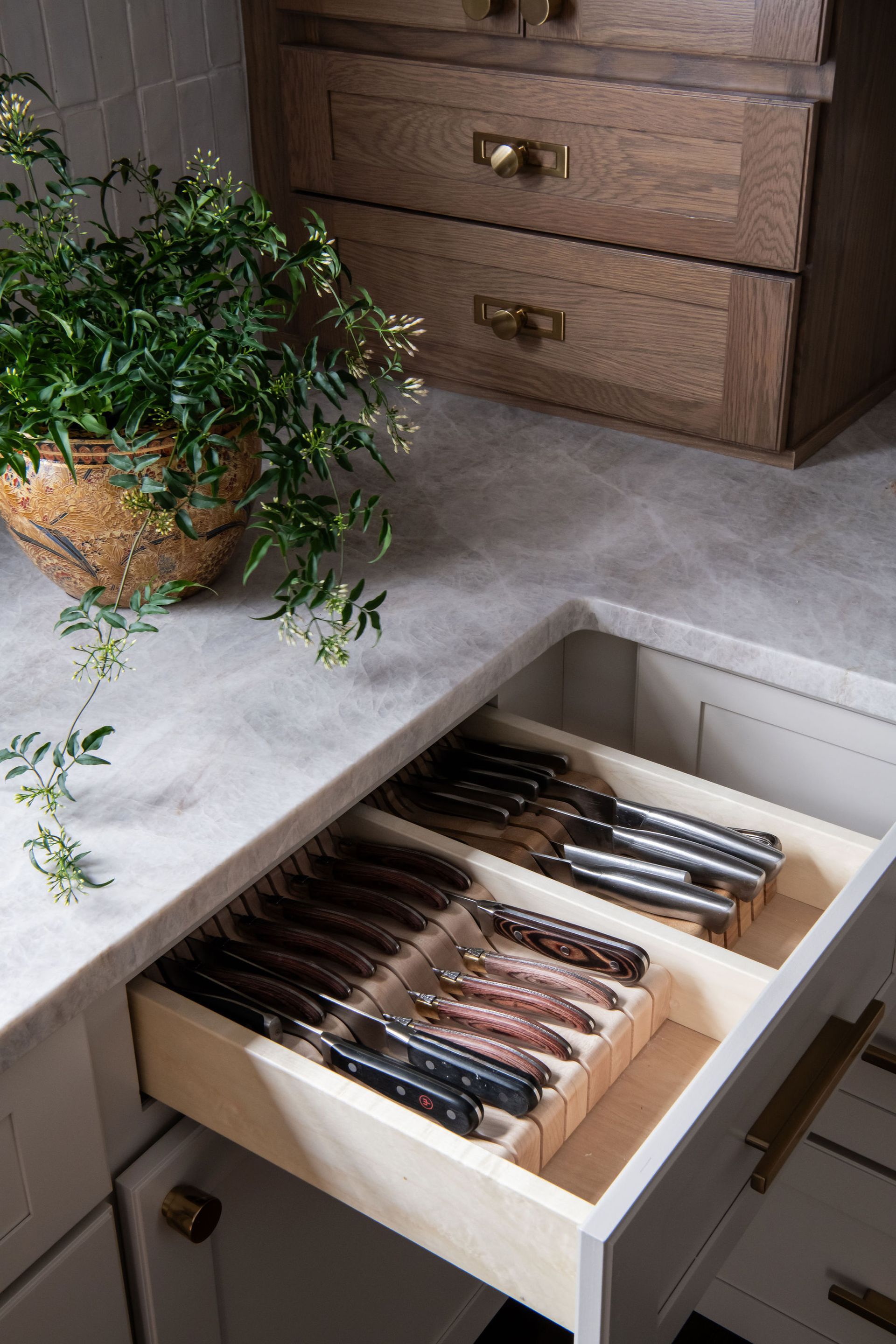 Cutlery drawer open, filled with knives and forks. Wooden cabinet, brass handles, on a countertop, with a plant.