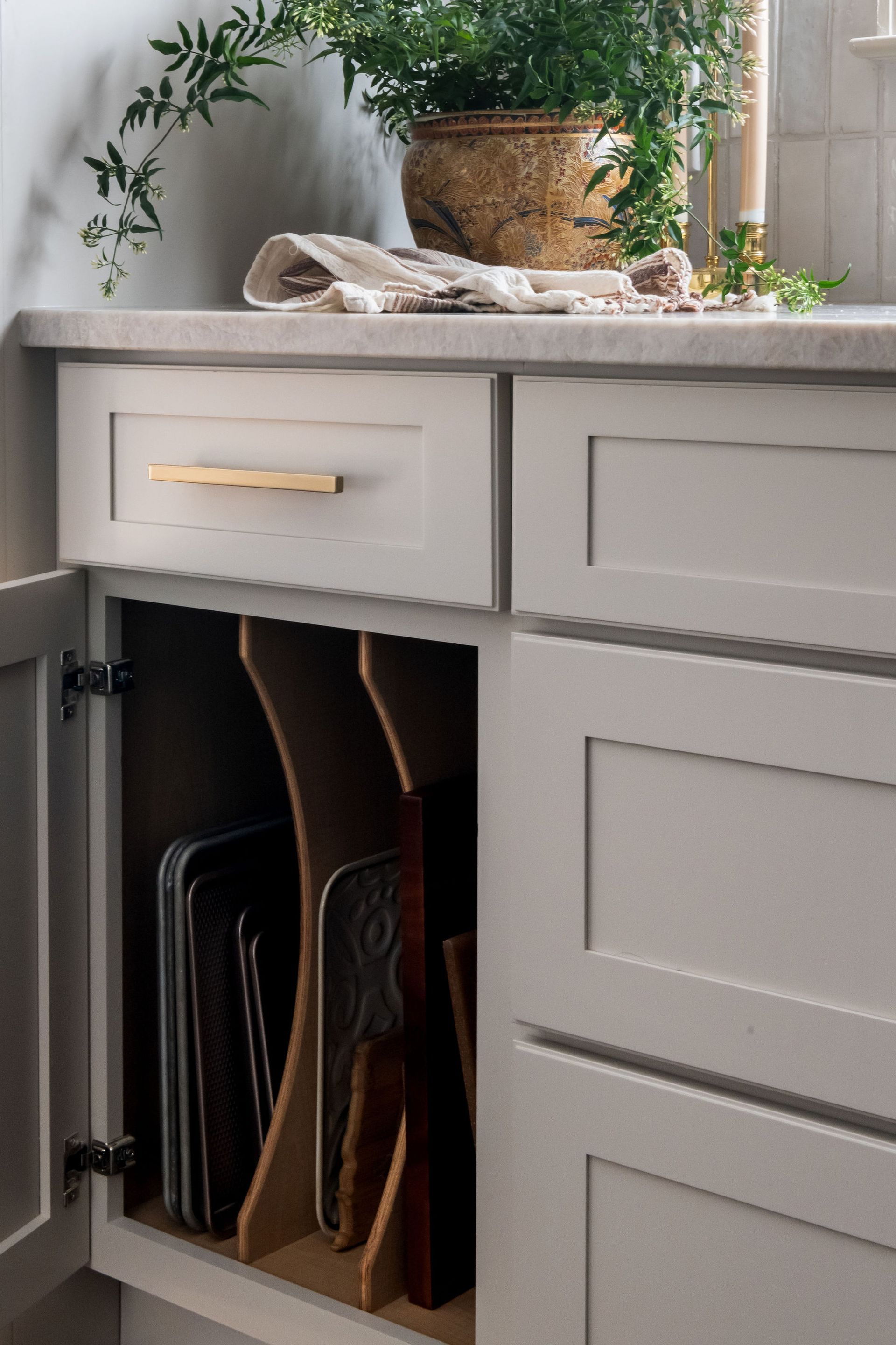 Open cabinet with baking sheets and cutting boards in a kitchen. Gray cabinets, light countertop, and a plant.