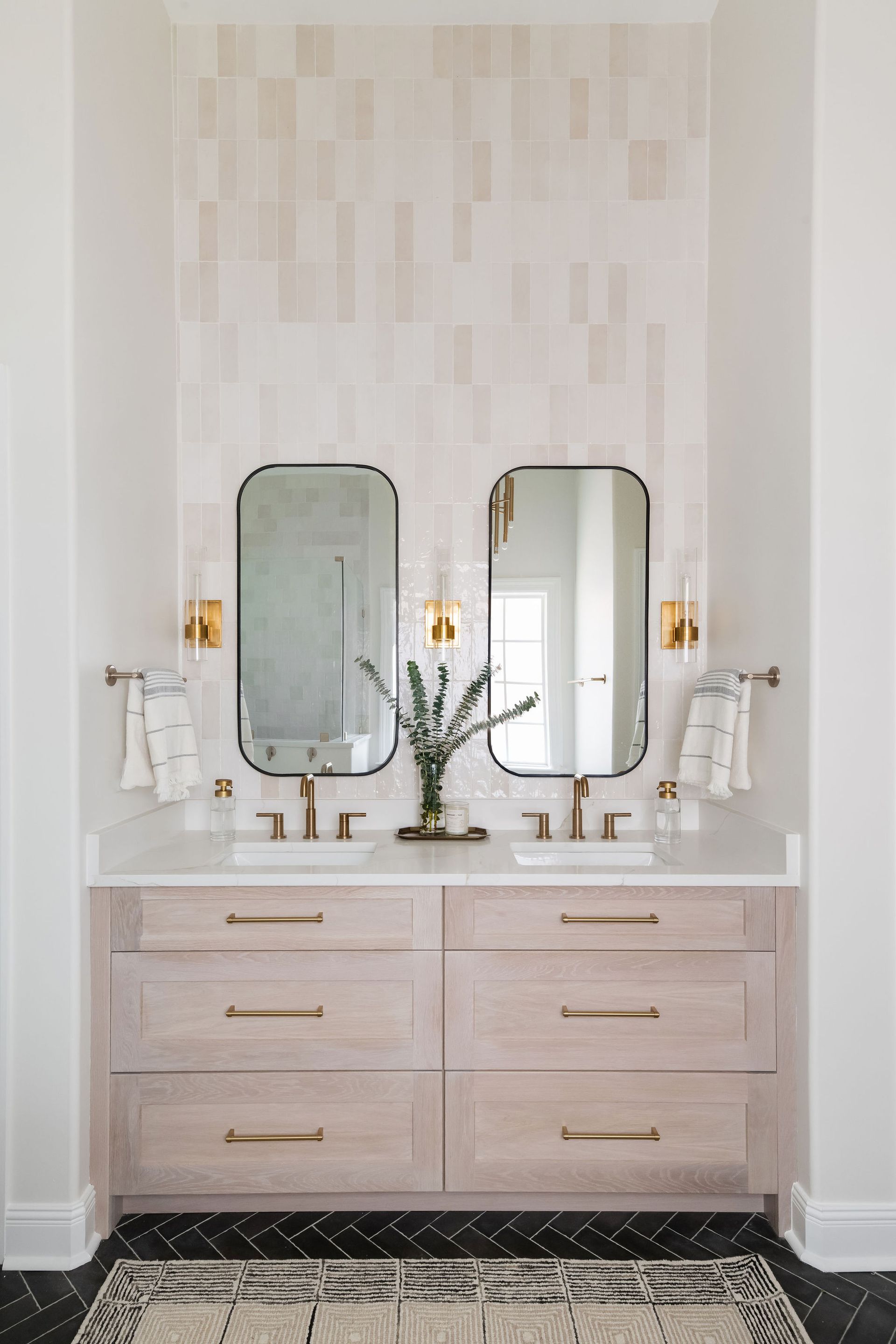 A modern bathroom vanity with two rectangular mirrors, gold fixtures, wood cabinetry, and white tile wall backsplash.