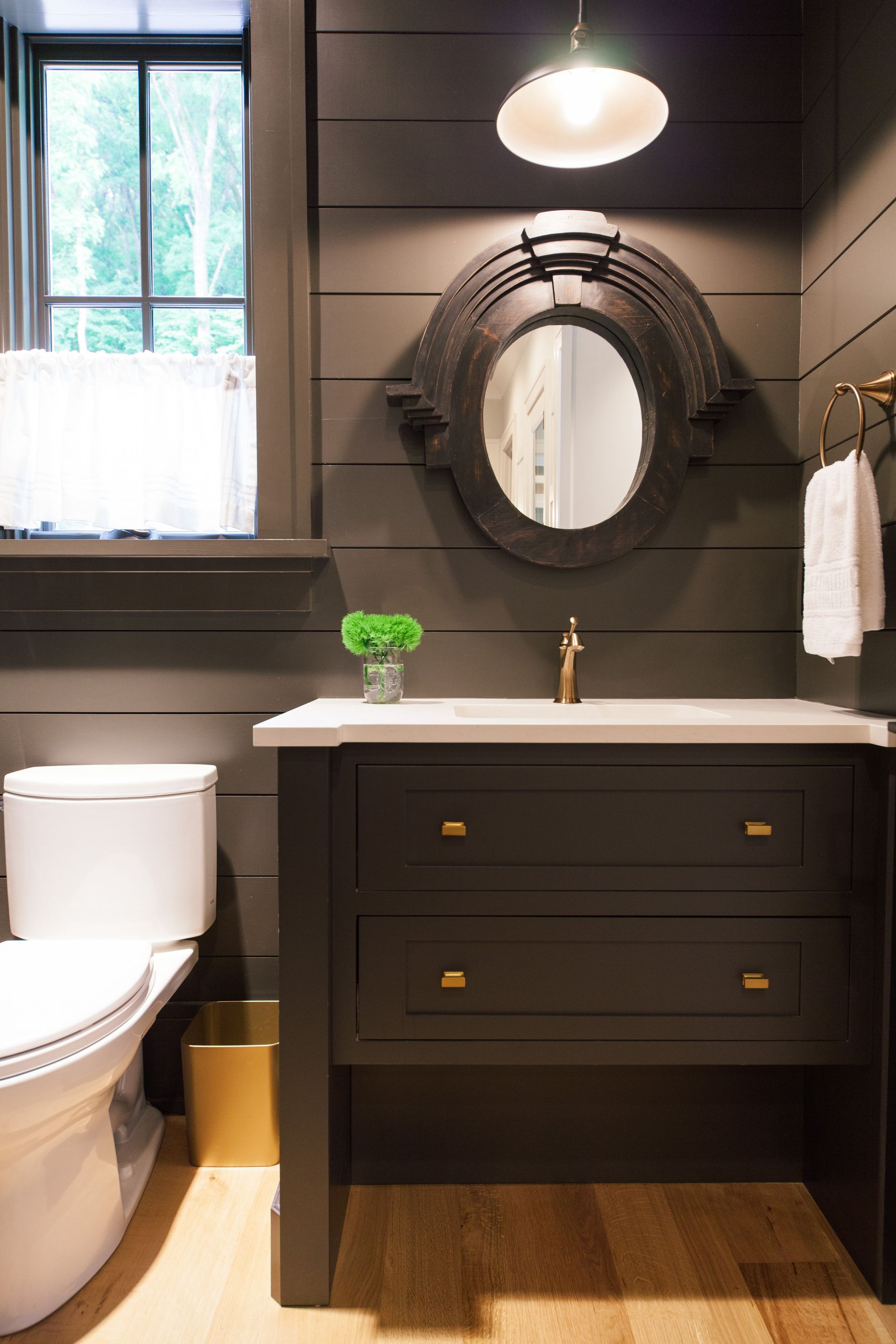 A dark-toned bathroom with shiplap walls, a wooden vanity, a white toilet, and an ornate oval mirror.