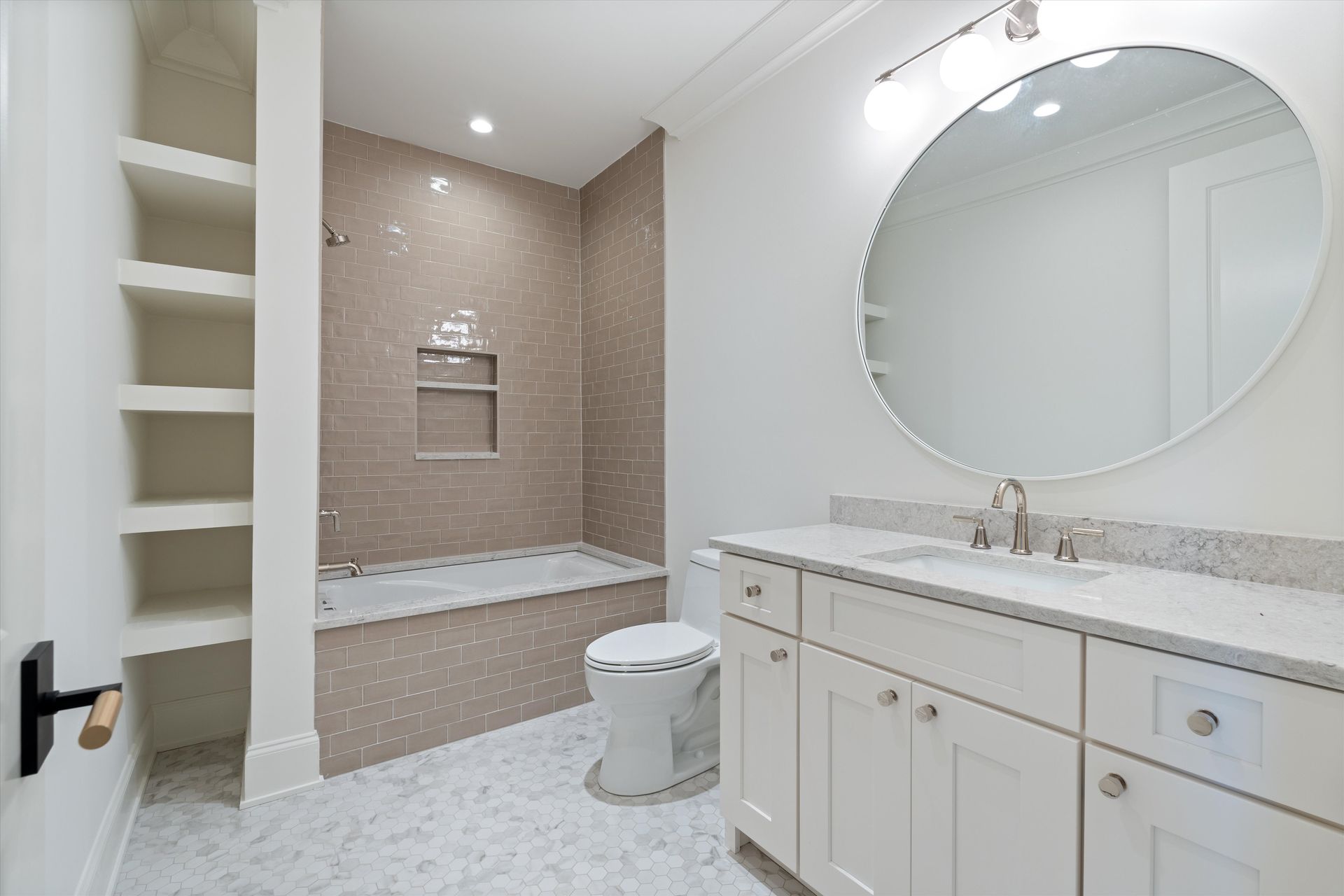 Modern bathroom with white vanity, oval mirror, toilet, and a bathtub alcove featuring beige tile and built-in shelving.